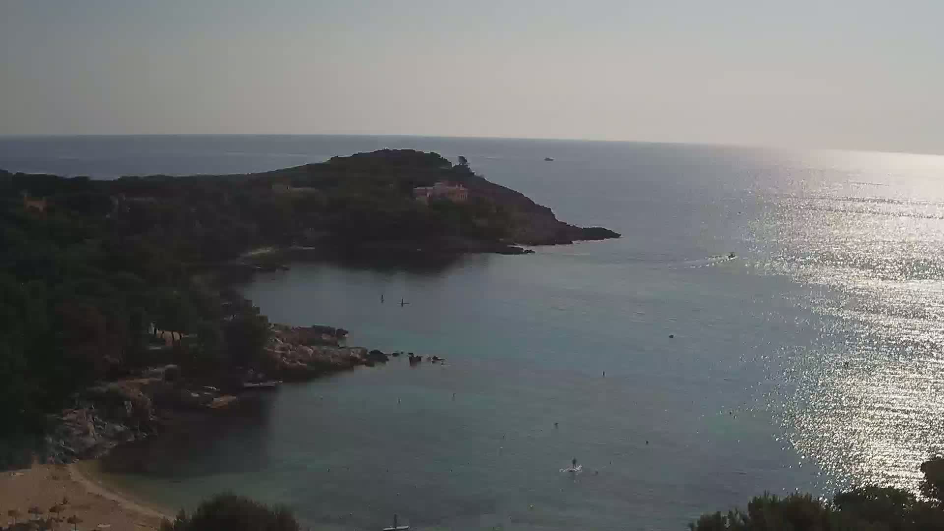 A calm, sunlit sea laps a rocky cove with a sandy beach, bordered by dark green vegetation and a small, tree-covered headland.