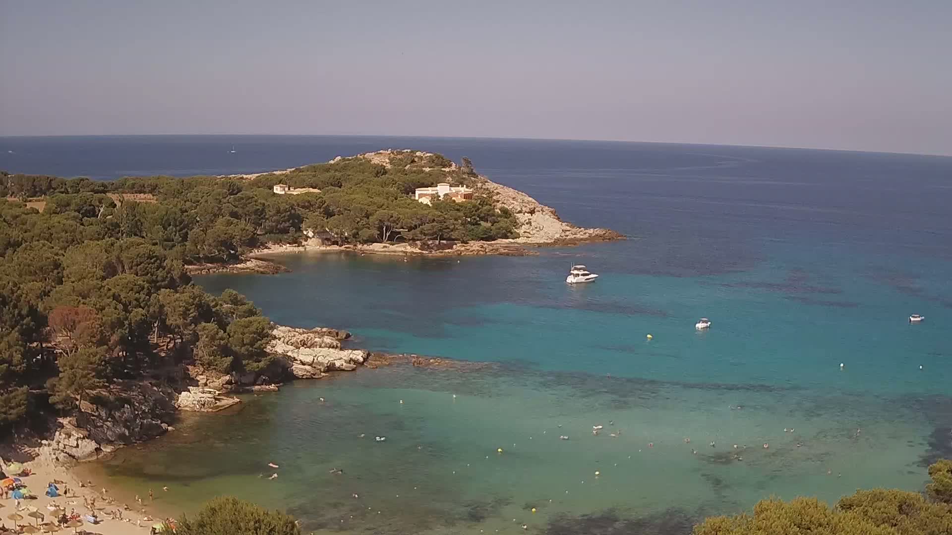 A tranquil cove with clear turquoise water, a sandy beach with bathers, and a small island covered in lush green vegetation under a clear sky.