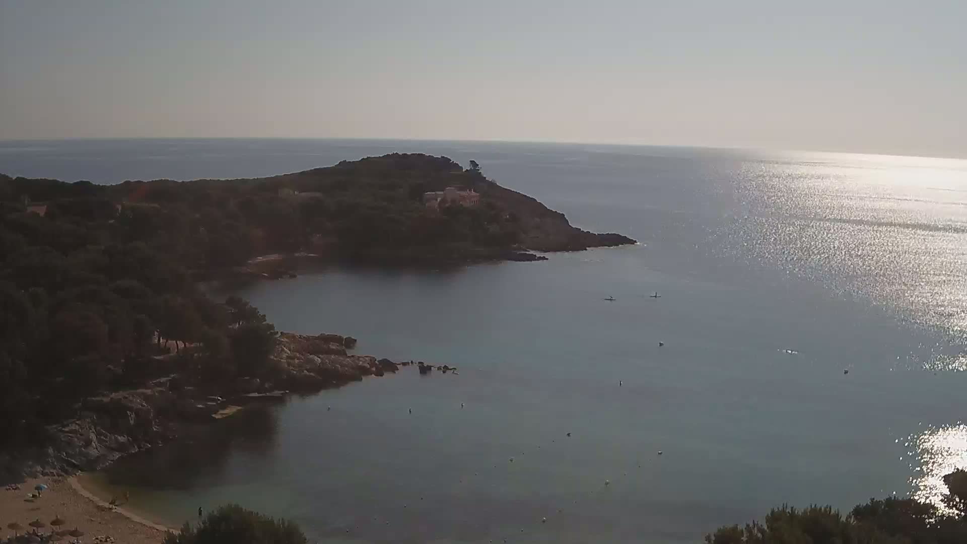 A secluded cove with a sandy beach and calm, sunlit water is nestled between rocky, tree-covered headlands.