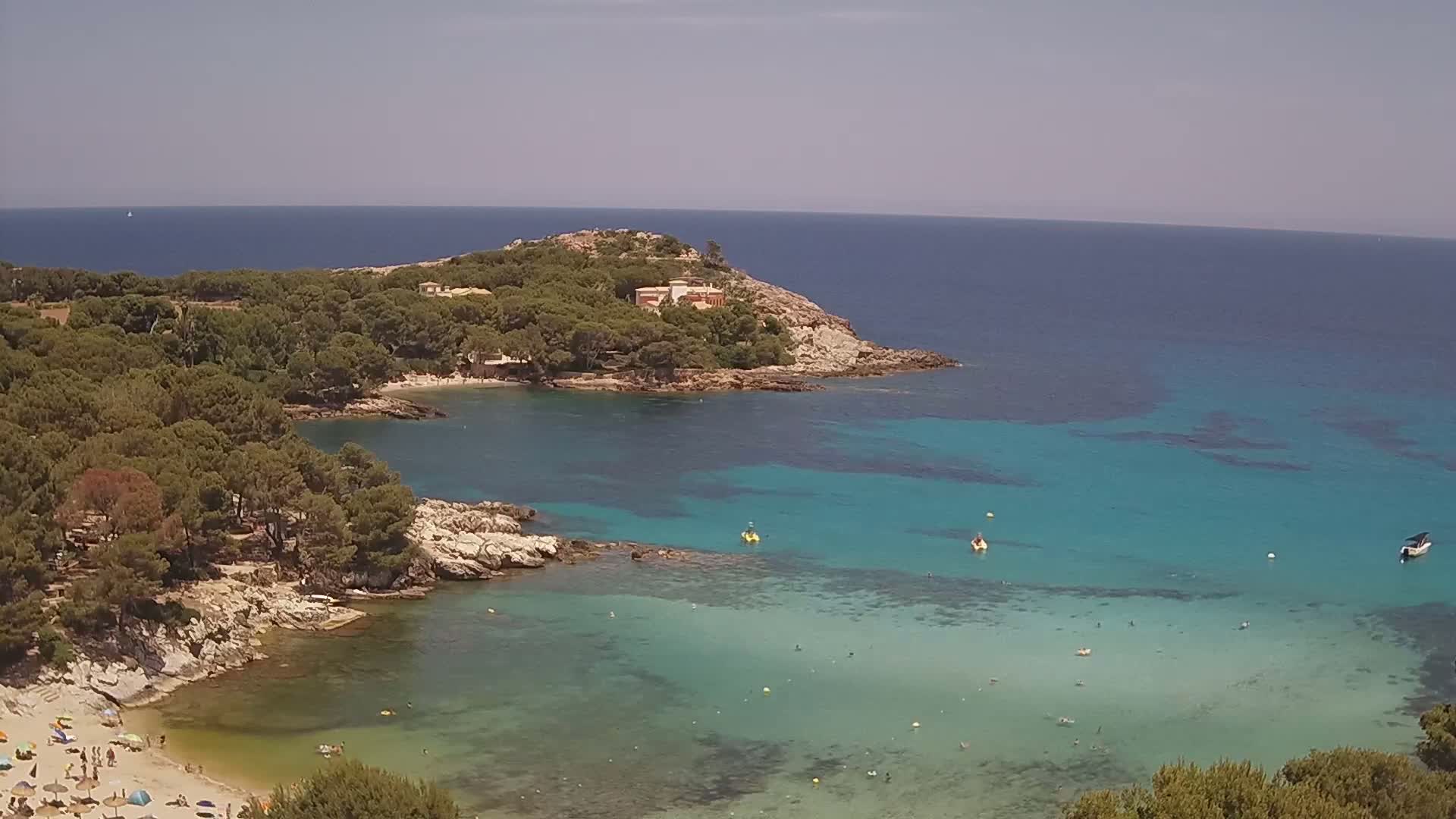 A tranquil cove with clear turquoise water, a sandy beach with people, and a small headland covered in pine trees under a sunny sky.