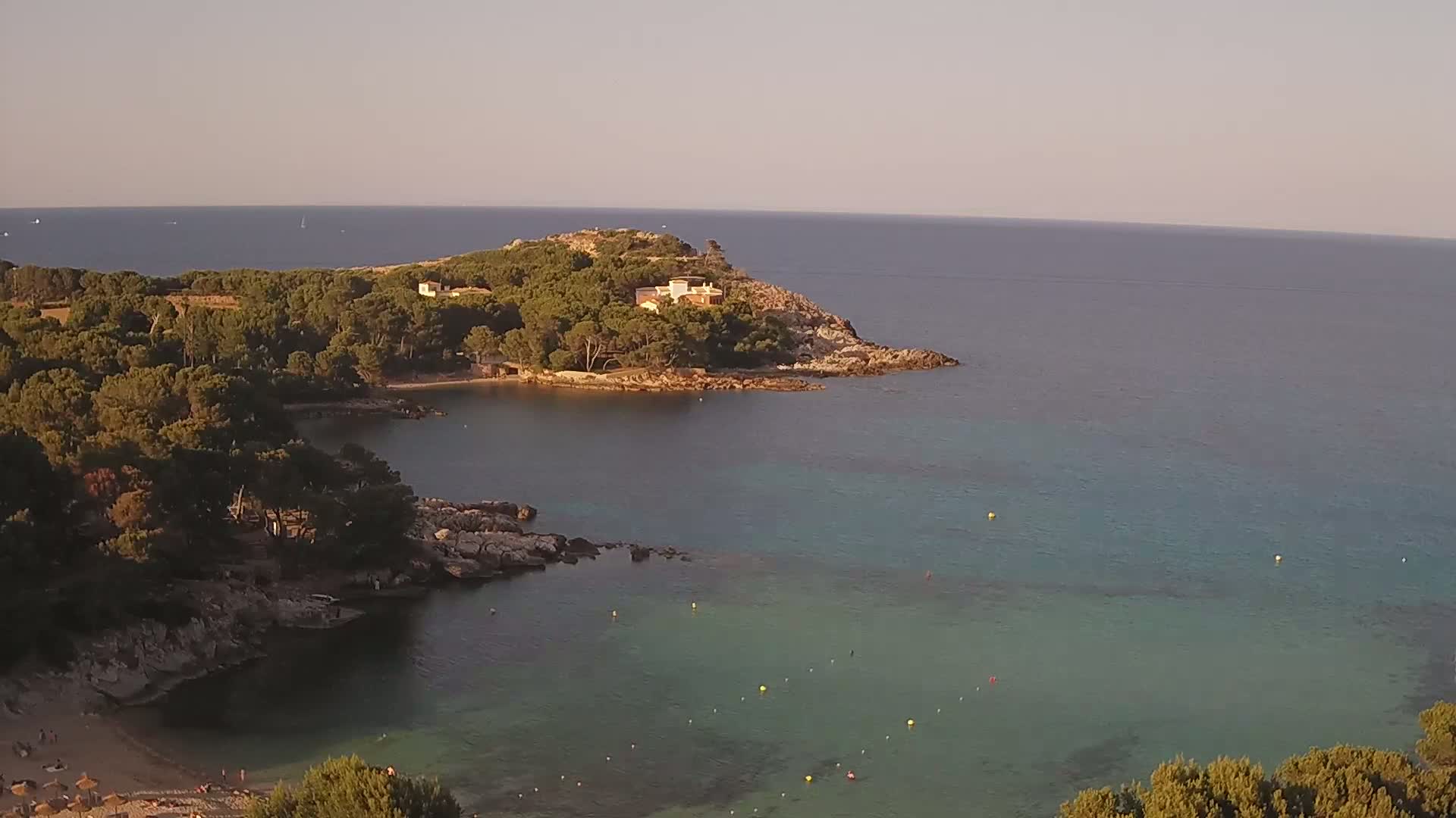 A tranquil cove with clear, teal water is nestled between a rocky shoreline and a verdant, tree-covered headland under a calm, light-colored sky.