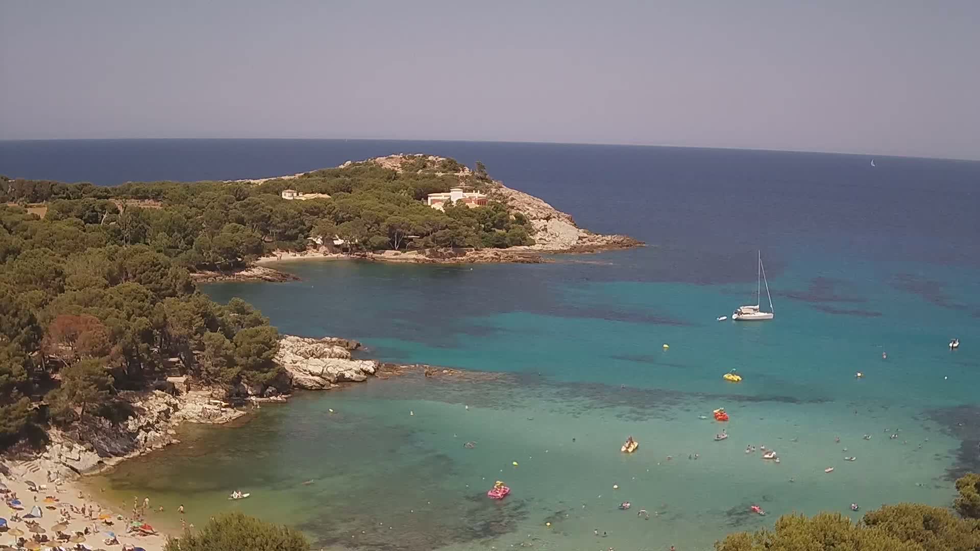A secluded cove with clear turquoise water, a sandy beach with bathers, and a small wooded peninsula under a clear sky.