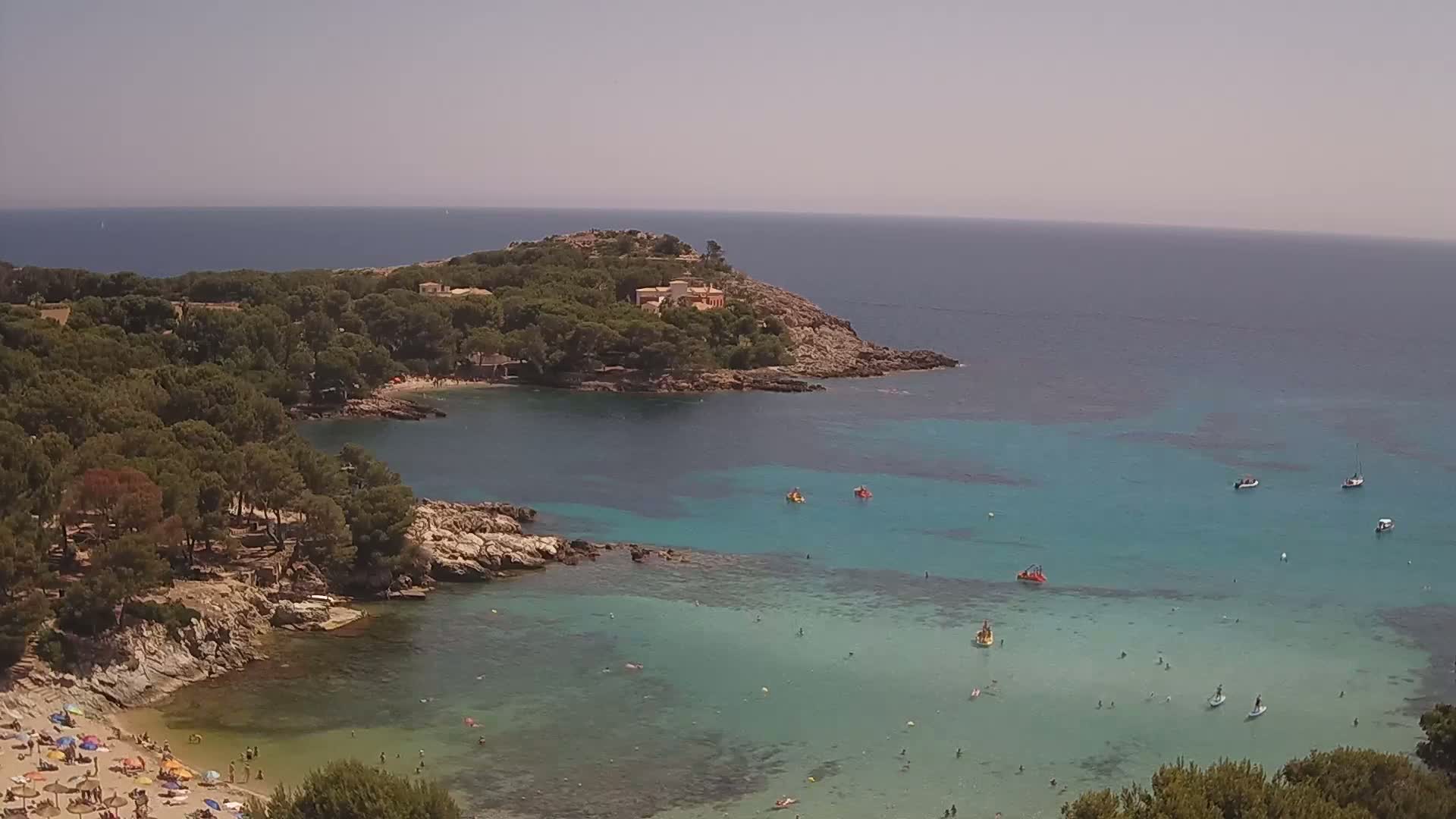 A clear, sunny day reveals a secluded cove with turquoise water, a sandy beach dotted with umbrellas, and a small rocky peninsula topped with buildings and lush greenery.