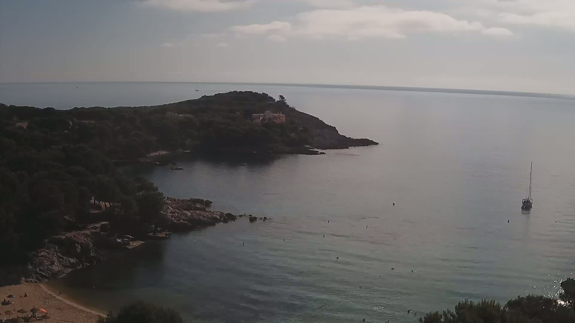 A tranquil coastal scene shows a sandy beach, rocky shoreline, calm sea with a sailboat, and a small house nestled on a tree-covered peninsula under a mostly cloudy sky.
