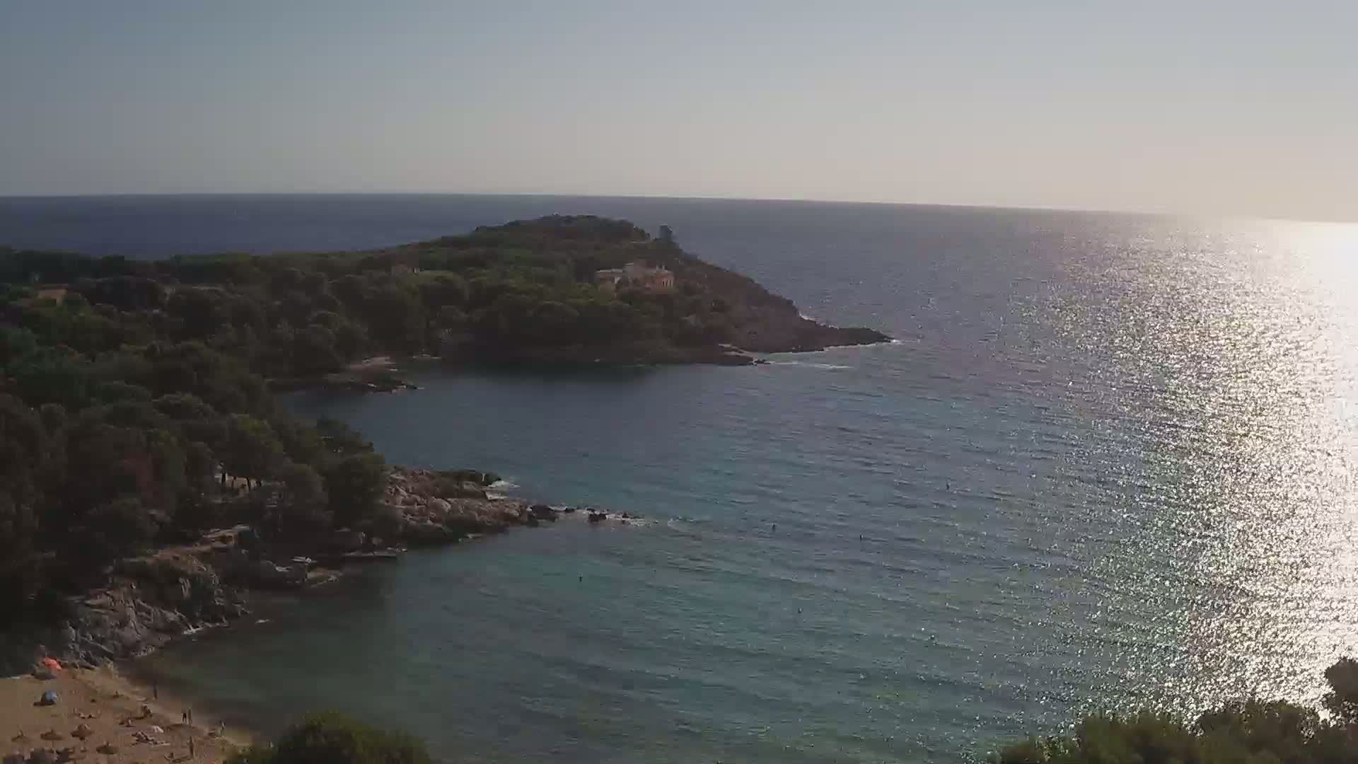 A sunny day shows a secluded cove with a sandy beach and calm, sparkling water surrounded by dark green trees and rocky shores.