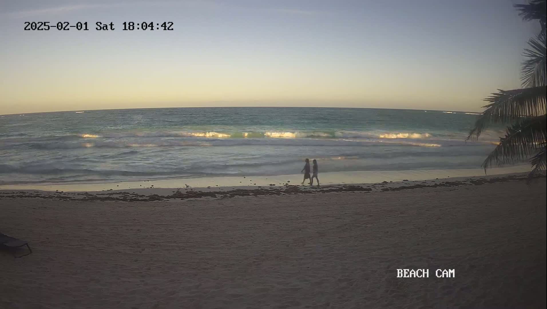 Two people walk along a sandy beach at sunset, with gentle waves lapping the shore under a clear sky.