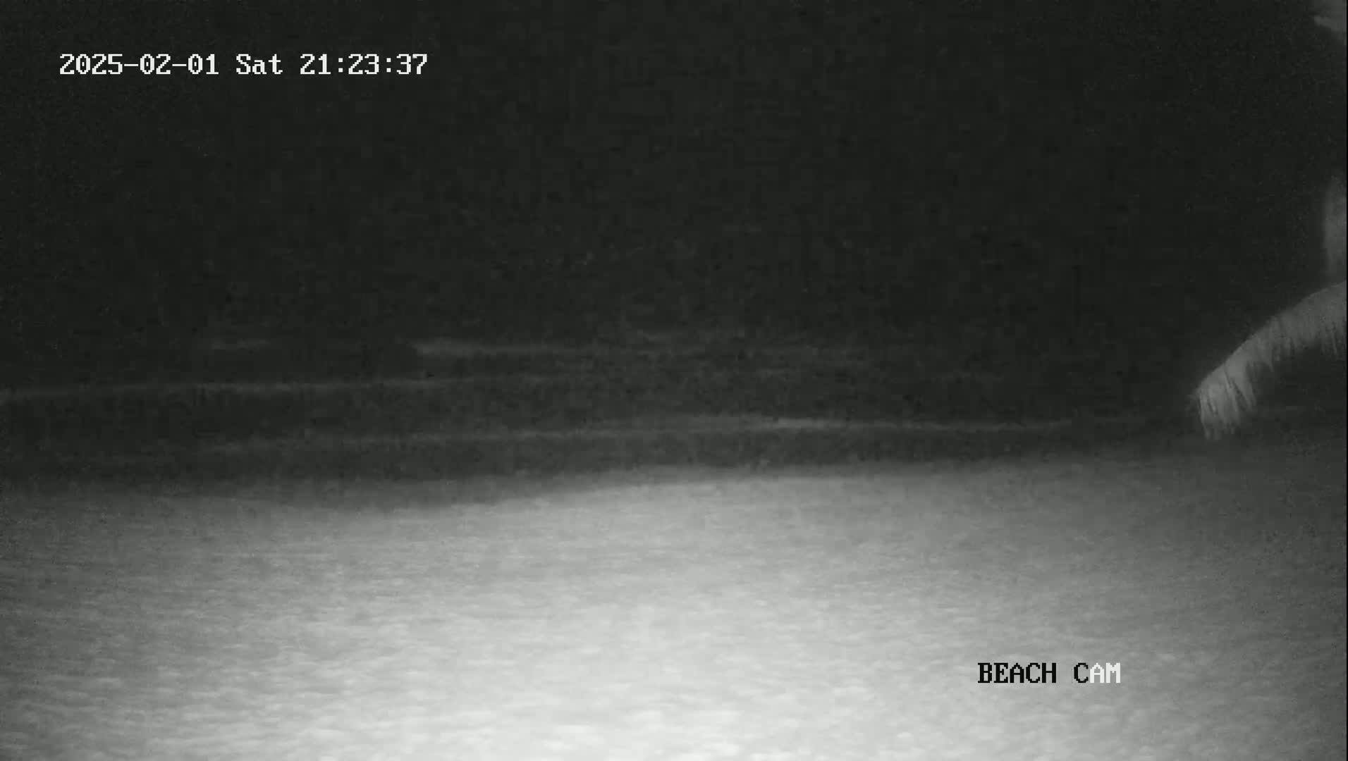 A nighttime beach scene shows dark water and a lighter colored sandy shore under a dark sky.