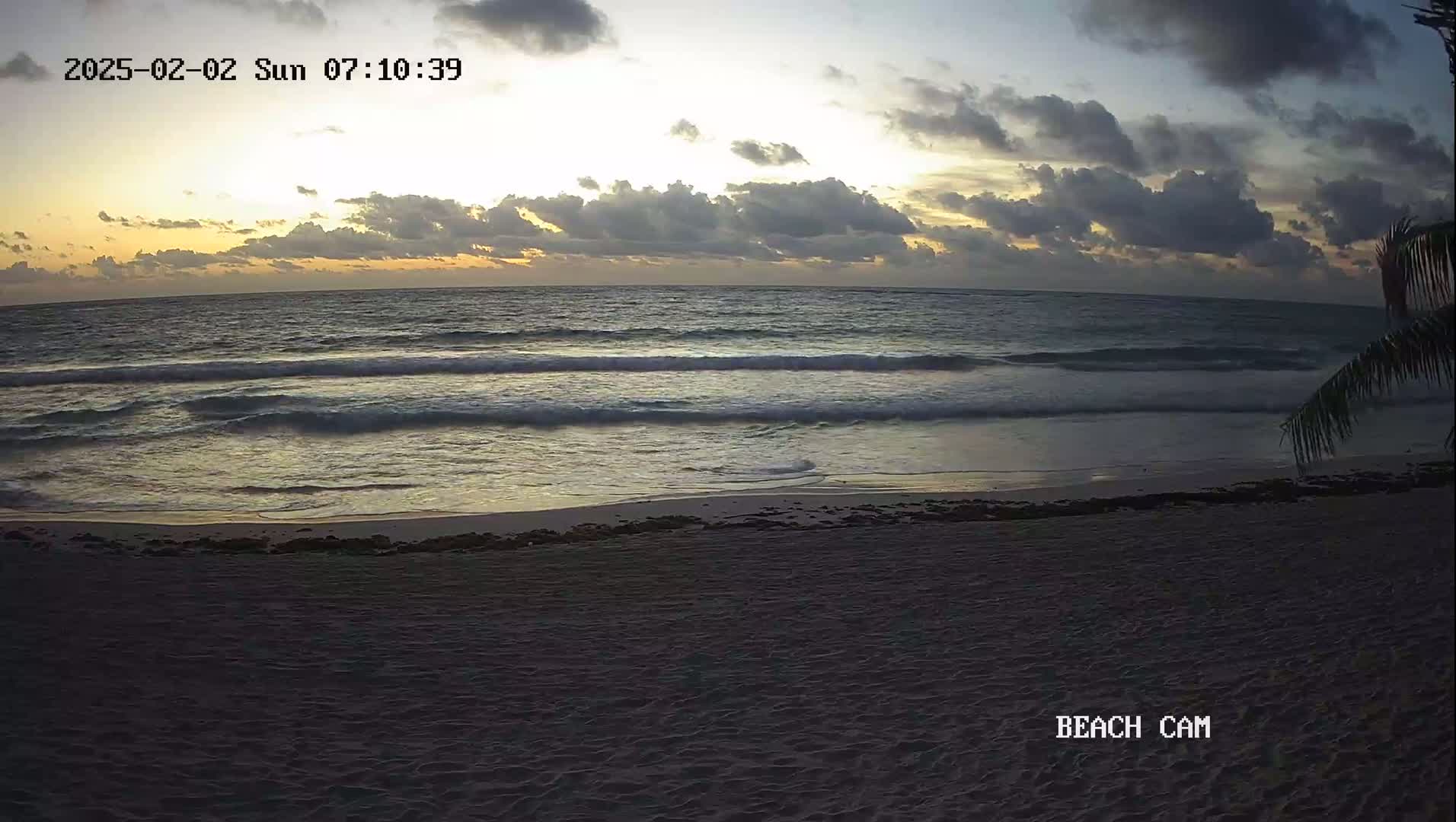 A sandy beach at sunrise shows gentle waves lapping the shore under a partly cloudy sky.