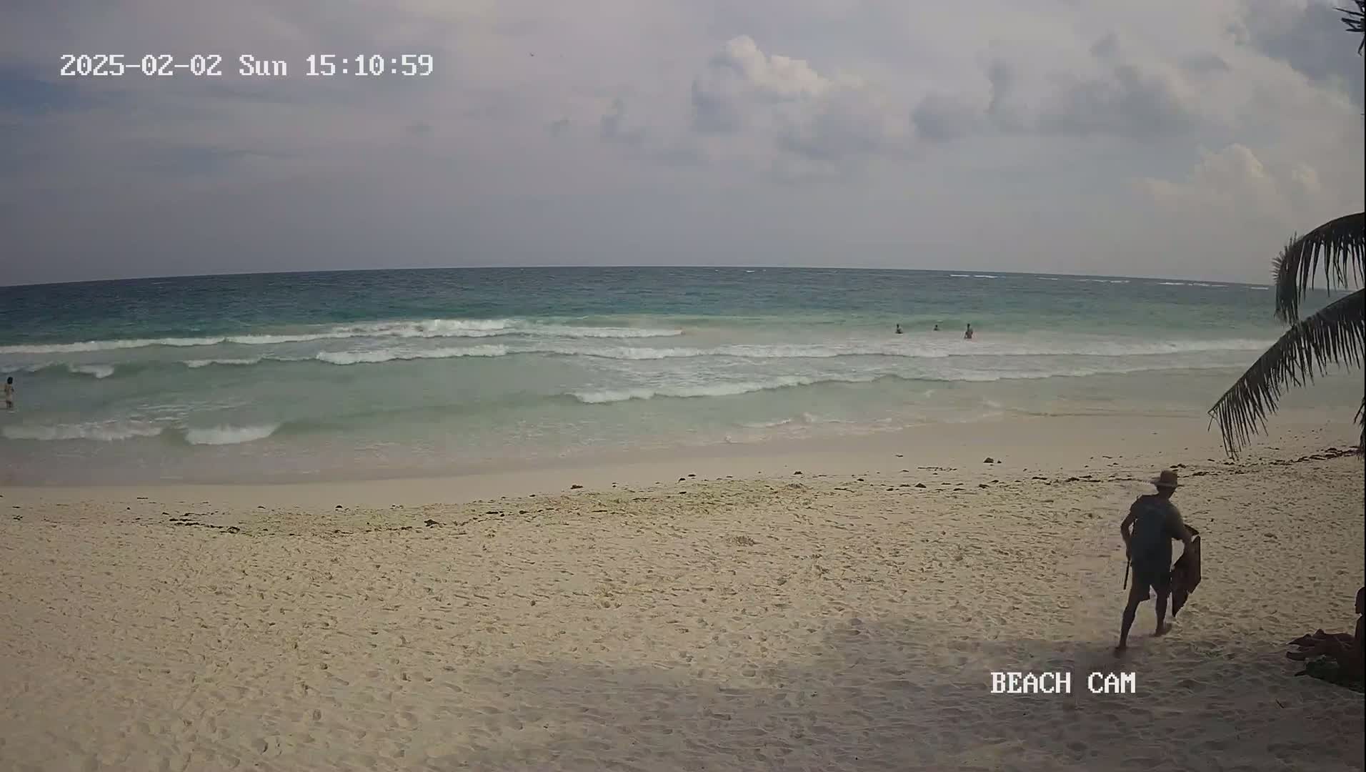 A sandy beach with several people in the water and one person walking on the sand under a cloudy sky.