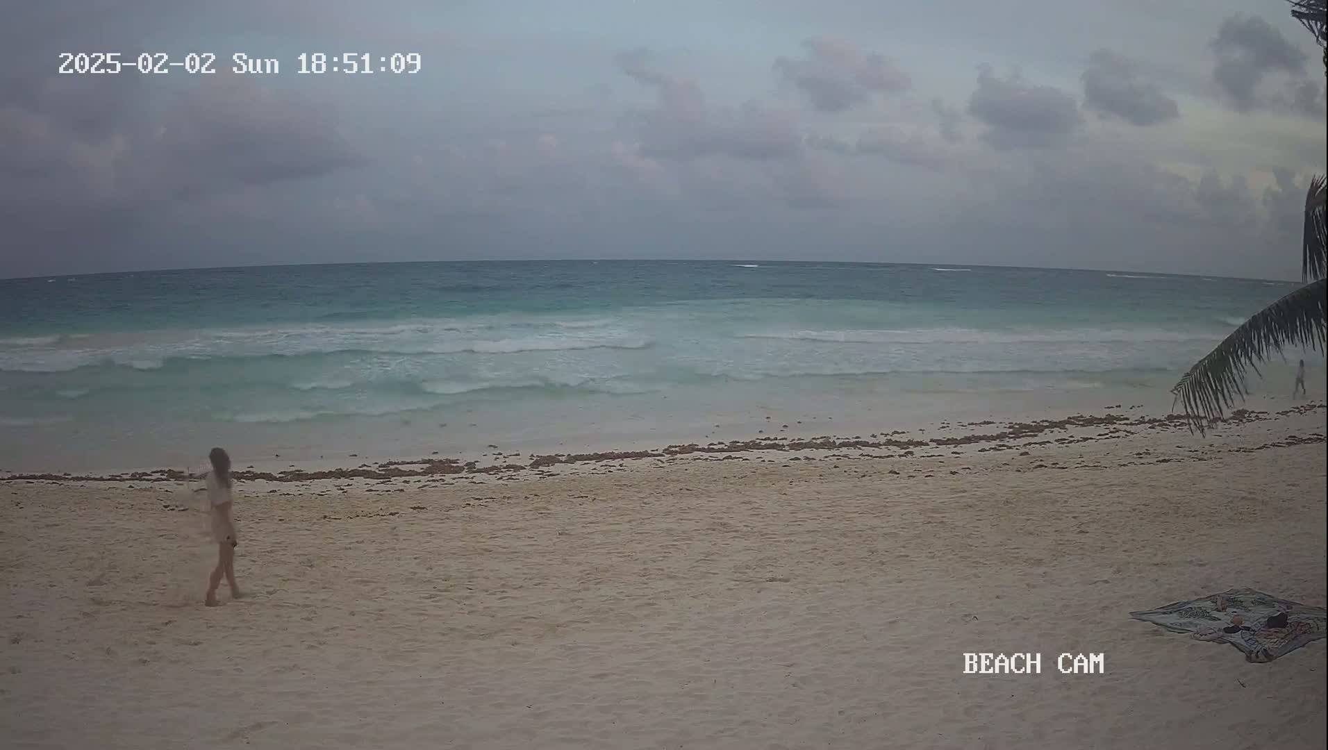 A woman walks on a sandy beach under an overcast sky, with moderately sized waves rolling into the shore.