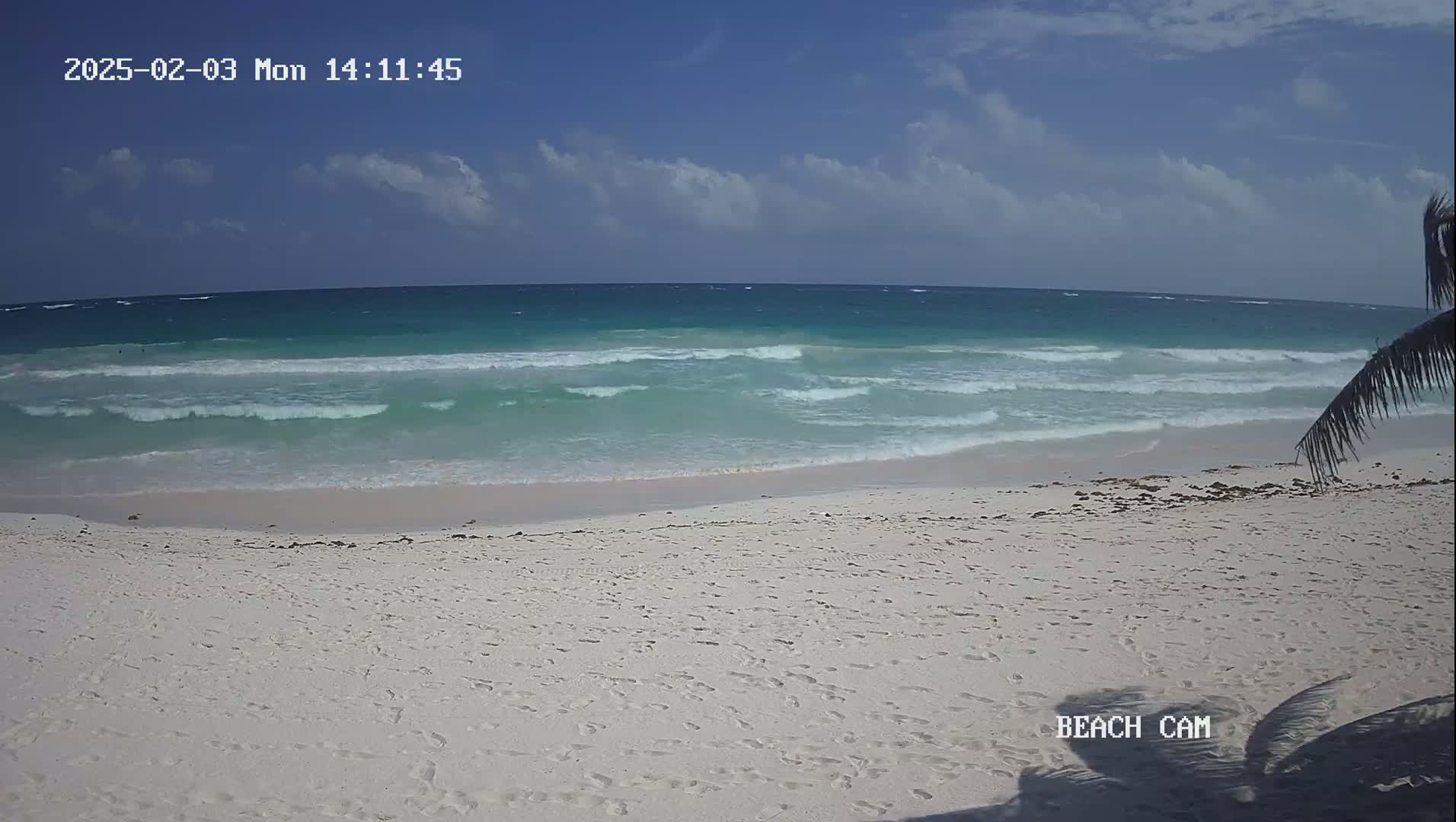 A white sandy beach with footprints is shown, with turquoise waves rolling onto the shore under a partly cloudy sky.