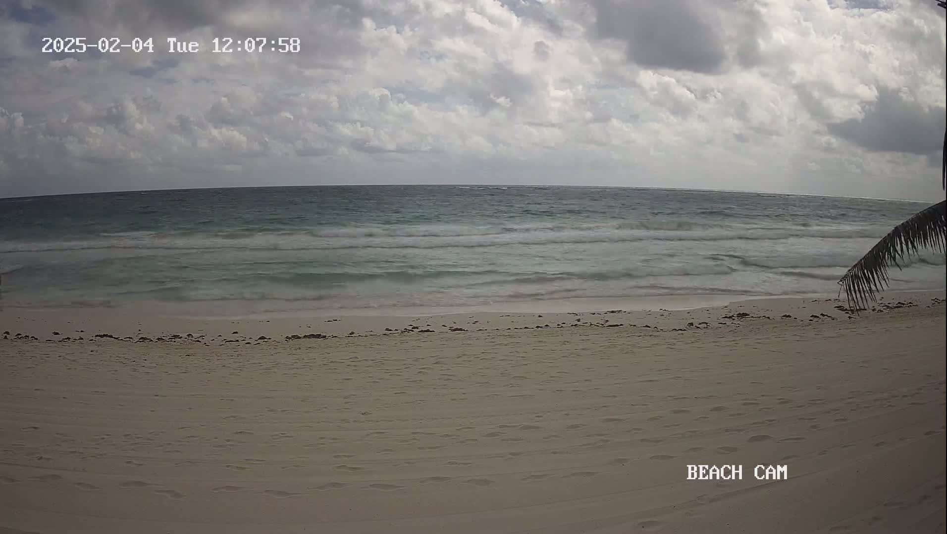 A mostly cloudy sky overlooks a wide, sandy beach with small waves lapping at the shore.