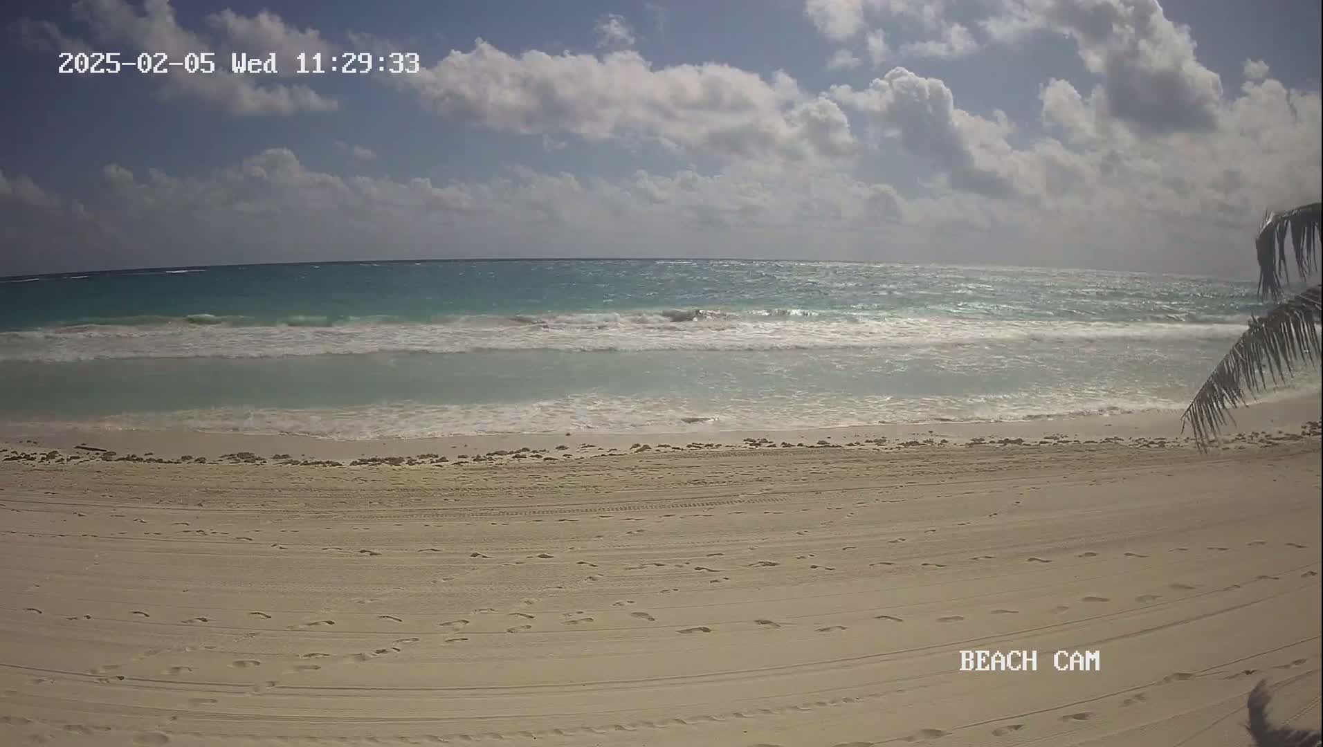 A sandy beach with footprints and tire tracks stretches toward a turquoise ocean with small waves under a partly cloudy sky.