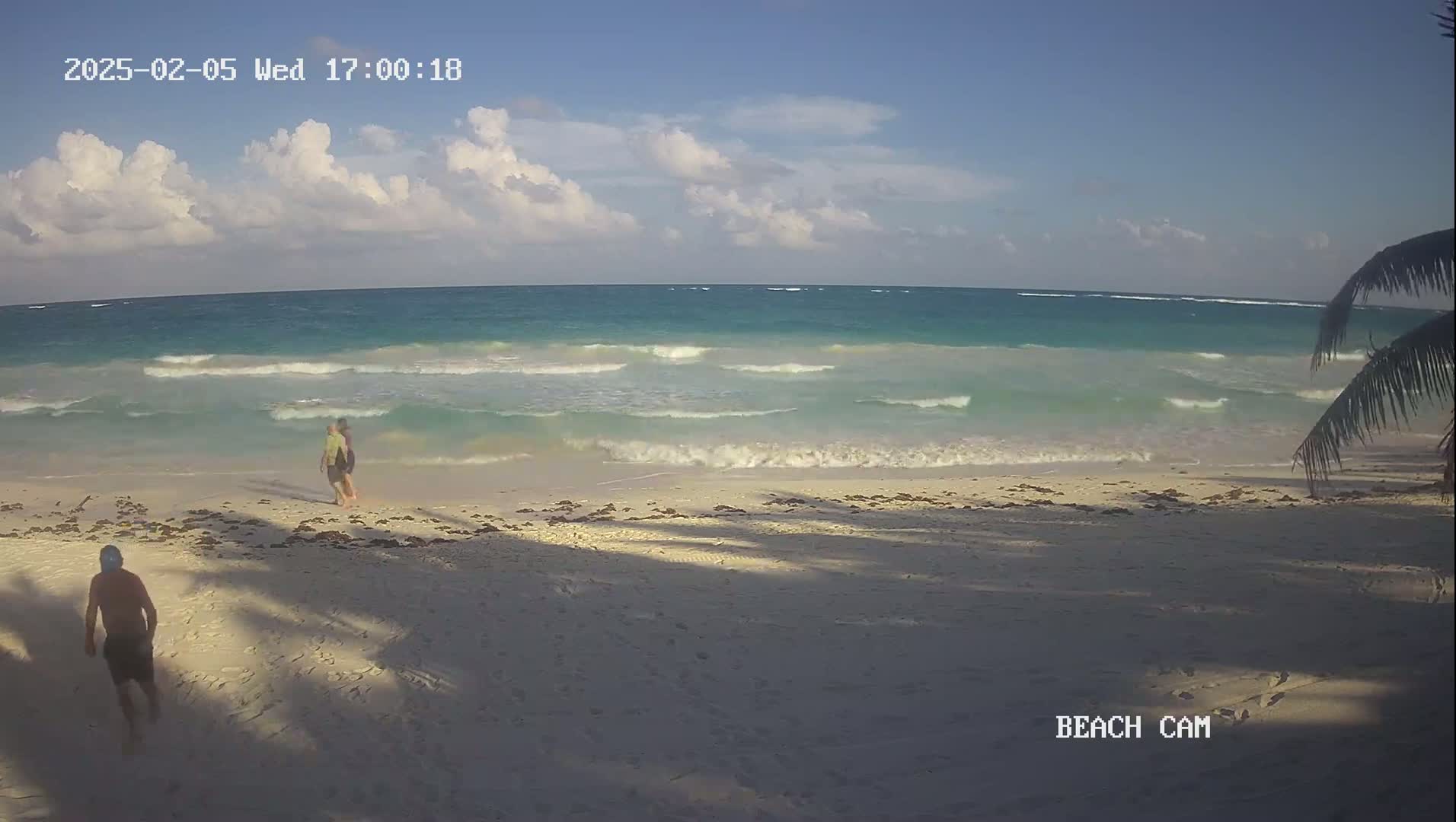 Three people walk along a sandy beach under a partly cloudy sky as small waves lap the shore.