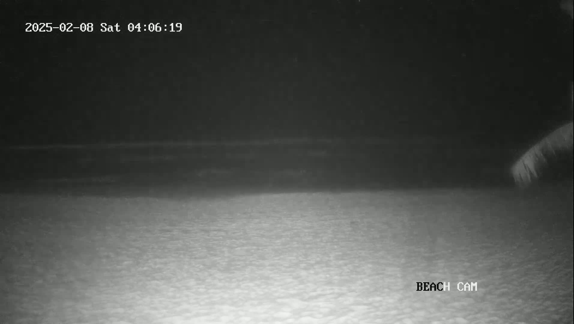 A nighttime beach scene shows dark water meeting a light-colored sandy shore under a dark sky.