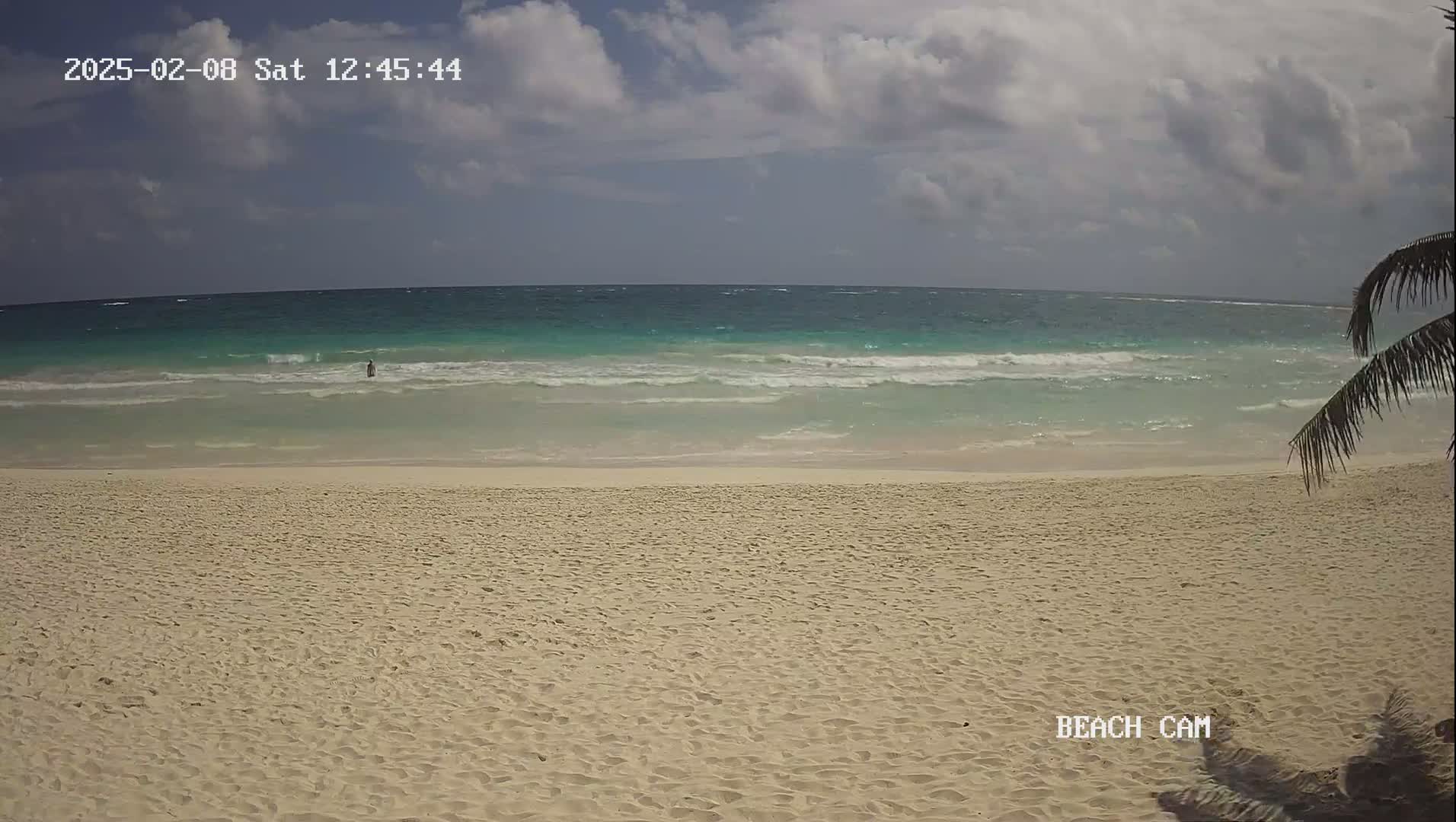 A mostly sunny day shows a wide sandy beach and turquoise ocean with small waves.