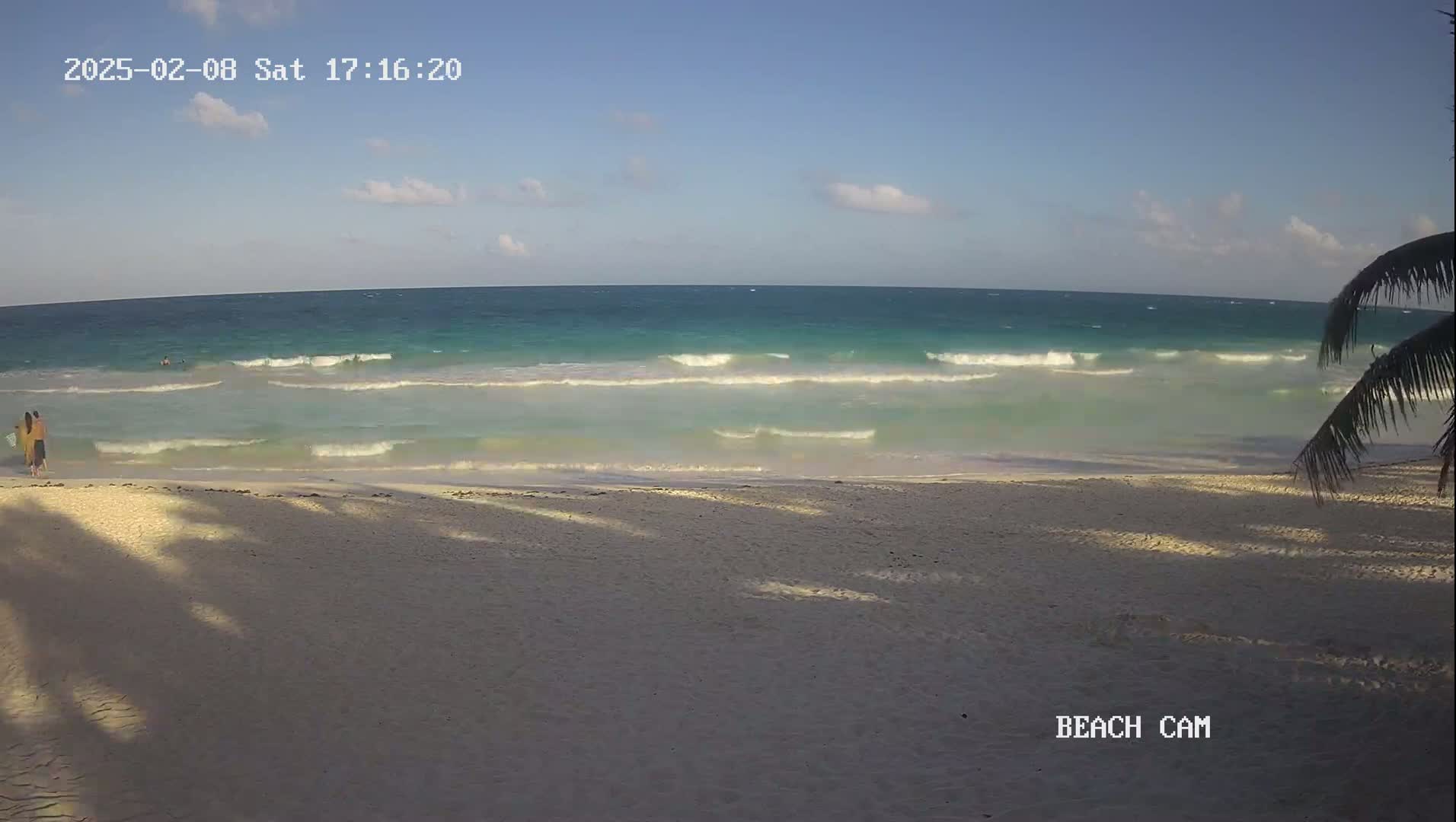 A sandy beach with gentle waves lapping the shore under a partly cloudy sky, with two people standing near the water's edge.