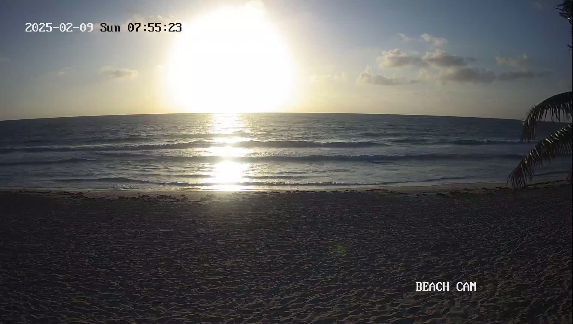 A sandy beach at sunrise with a calm ocean and a few clouds in the sky.