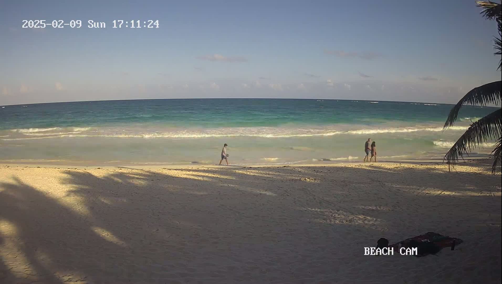 A sandy beach with gentle waves on a clear day, a few people walking on the shore, and palm tree shadows cast on the sand.