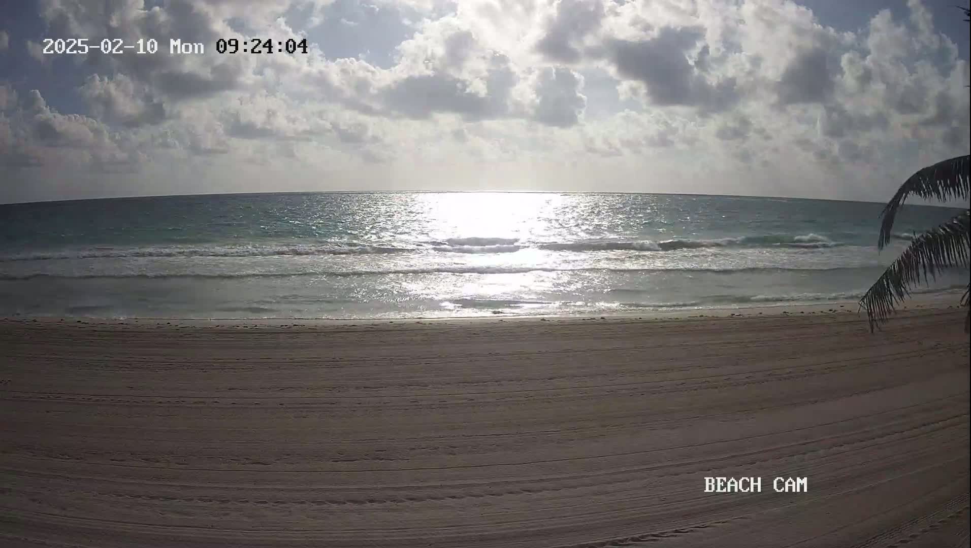 A partly cloudy sky shines down on a sandy beach with gentle waves lapping the shore.