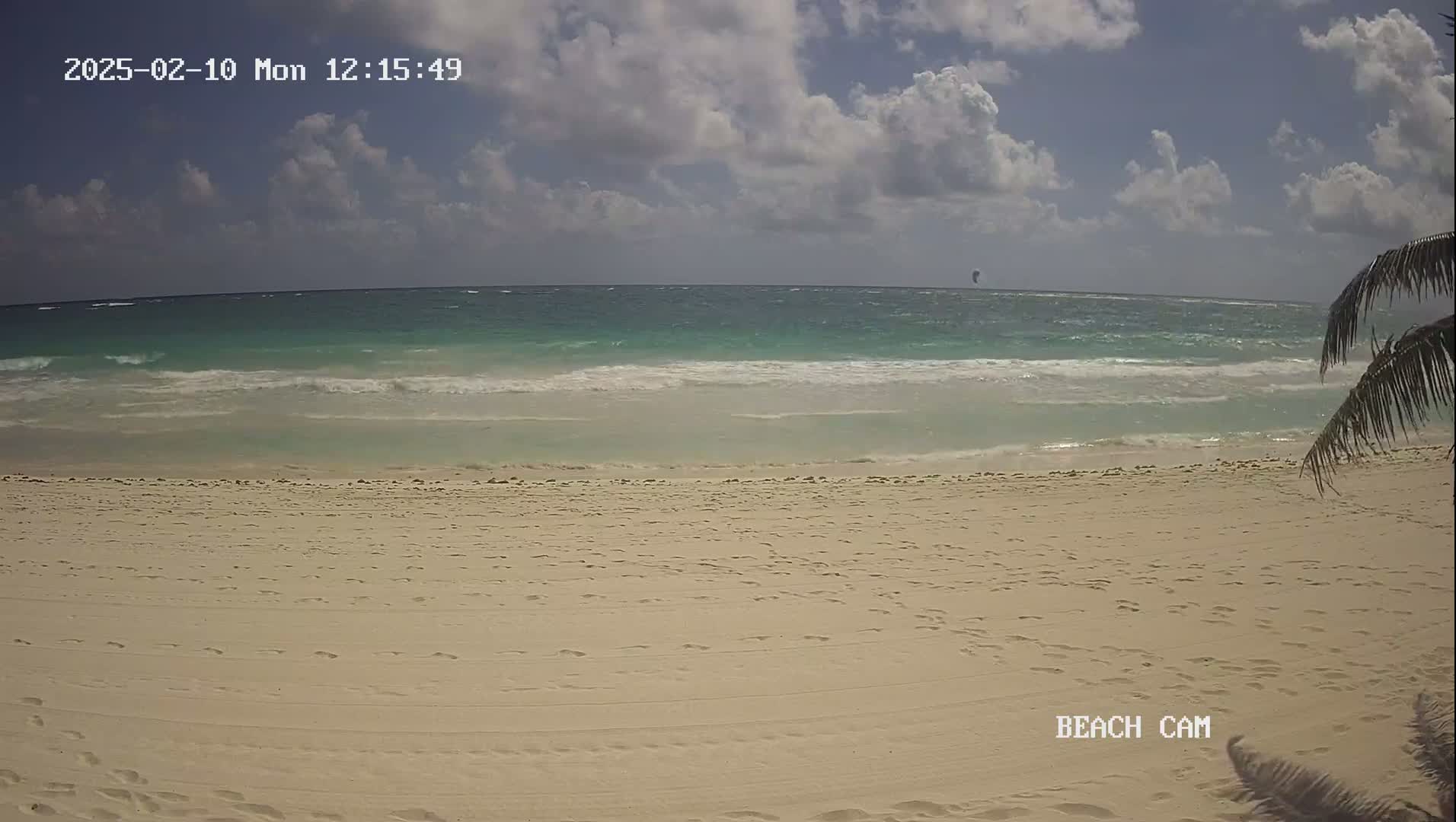 A partly cloudy day shows a sandy beach with small waves lapping the shore, and a lone kitesurfer in the distance.