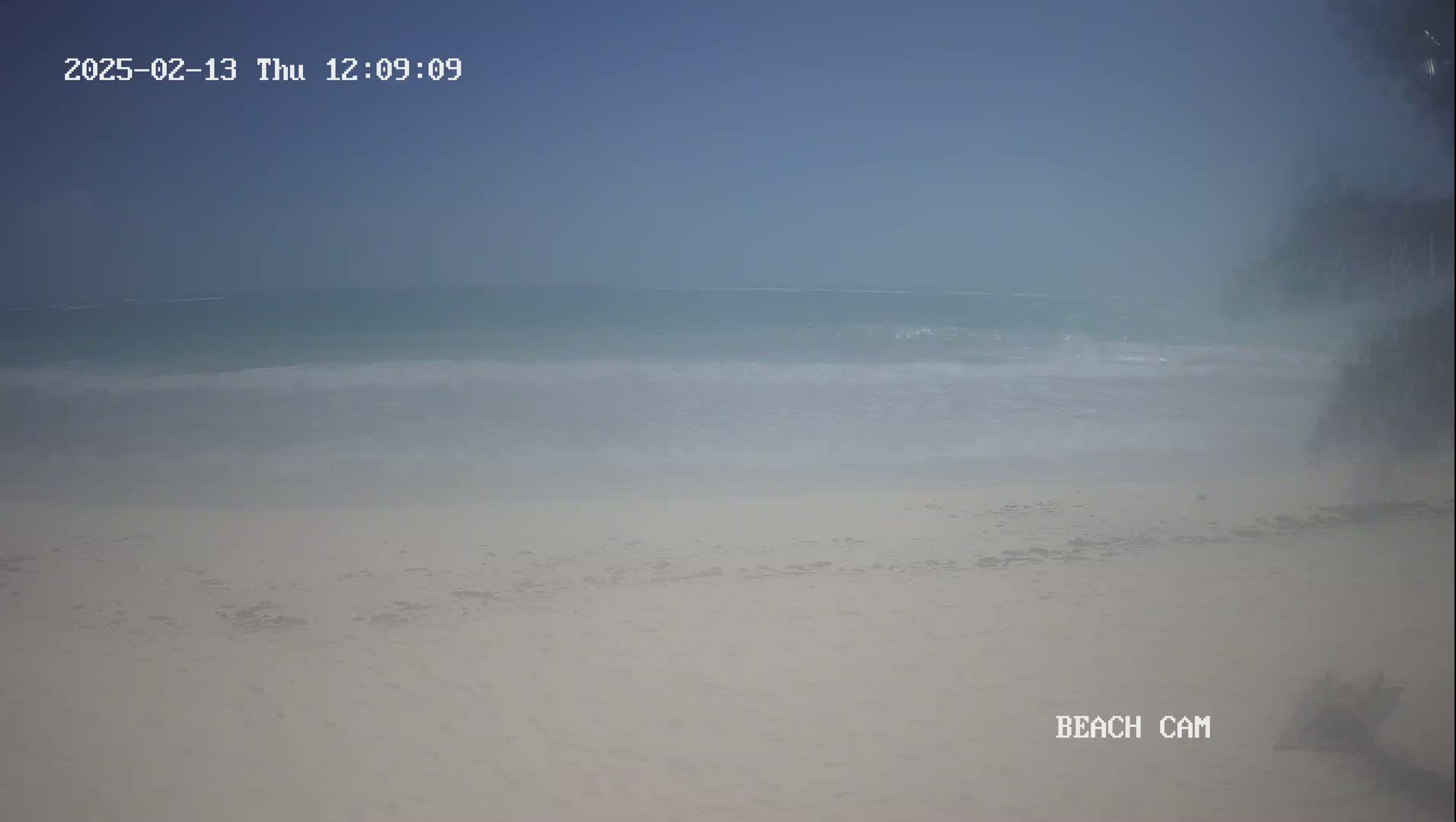 A hazy, light-blue ocean meets a wide, pale sand beach under a clear sky.