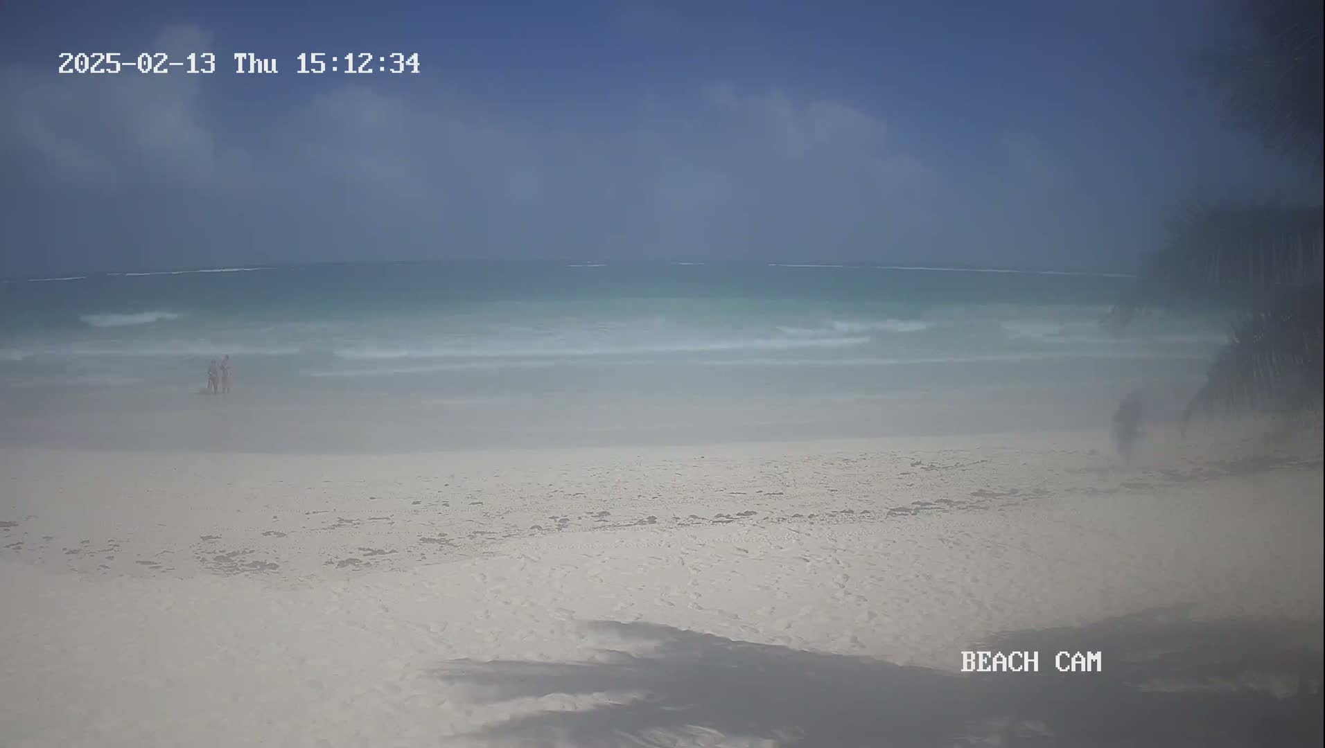 A hazy beach scene shows a wide expanse of light-colored sand and a calm, turquoise ocean under a mostly clear sky.