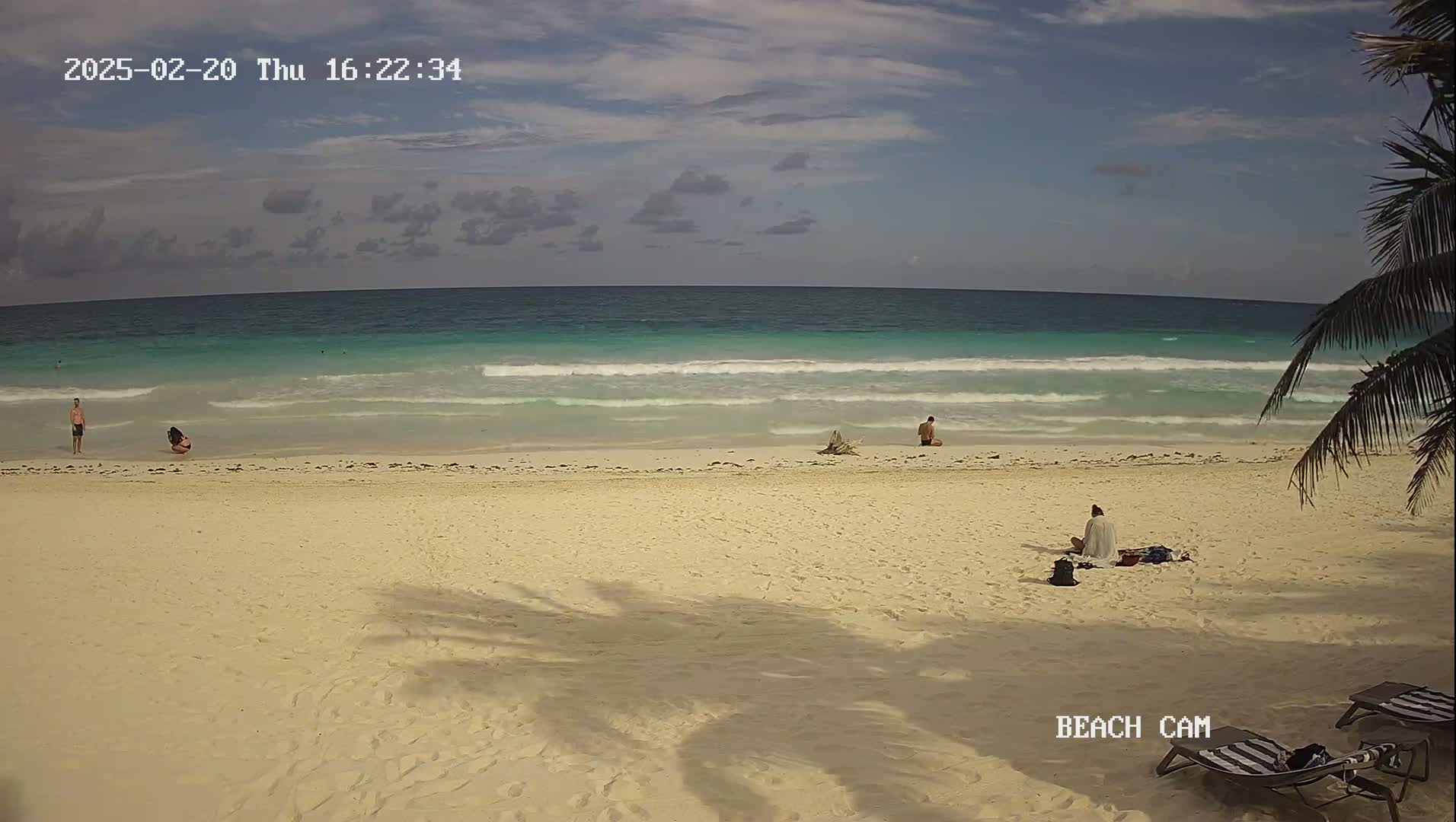 A mostly sunny beach scene shows a light-colored sandy beach with a few people and beach chairs, along with a calm turquoise ocean.