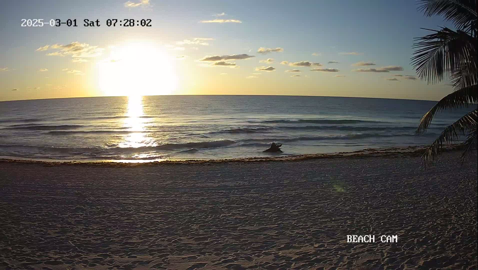 A sandy beach at sunrise shows calm ocean waves lapping the shore under a partly cloudy sky.