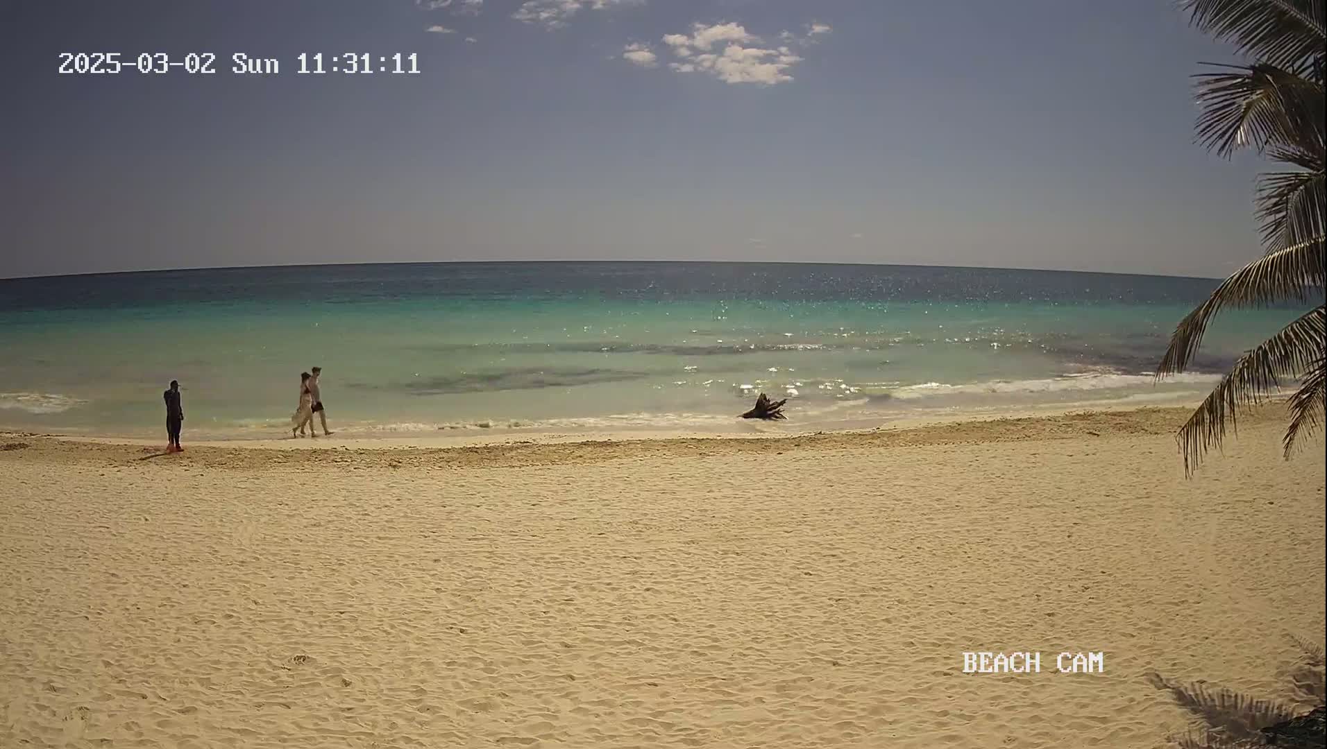 A sandy beach with turquoise water under a mostly clear sky shows three people walking near the water's edge, with one person standing further away.