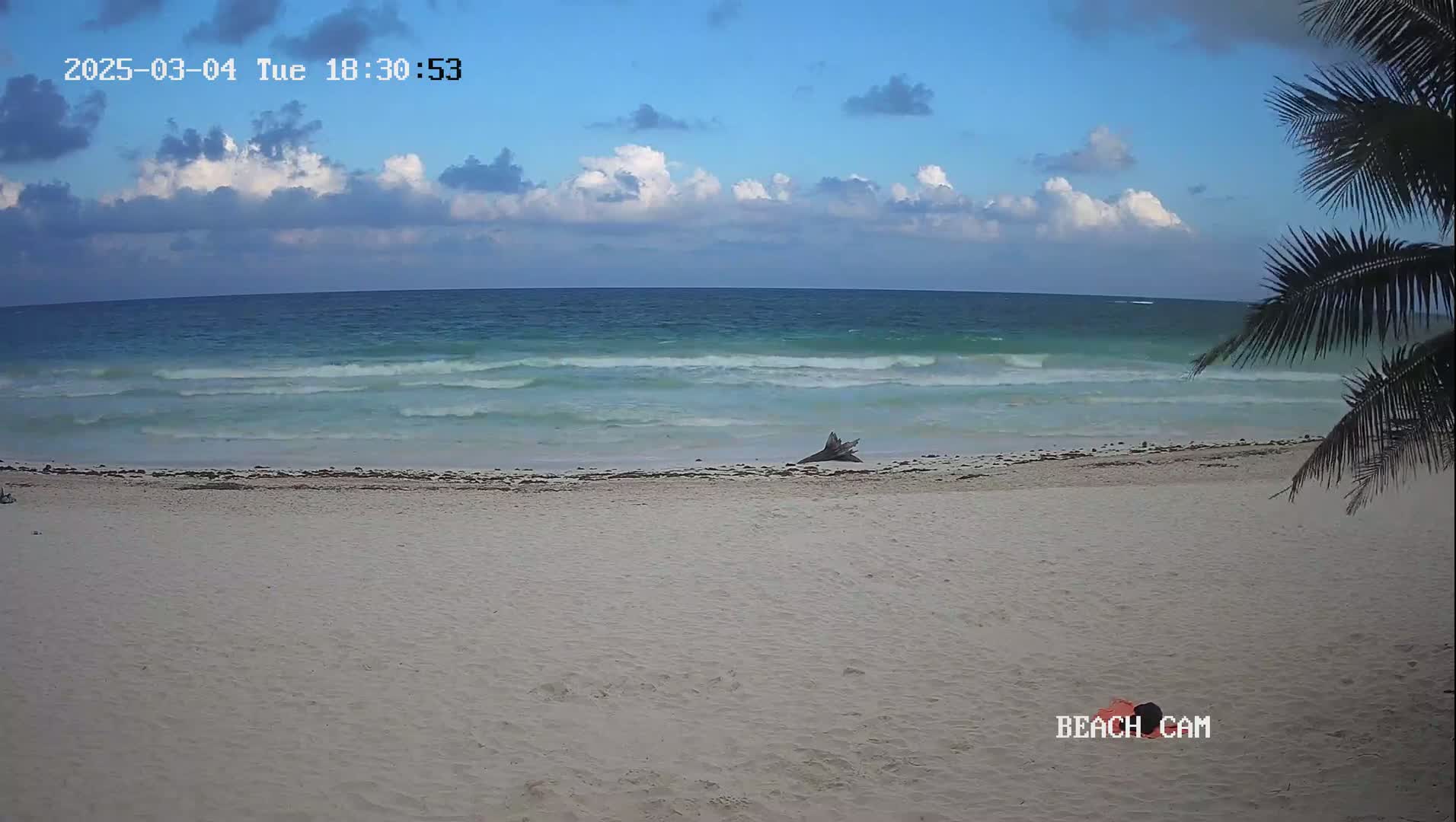 A sandy beach with light-colored sand and small waves on a partly cloudy day.