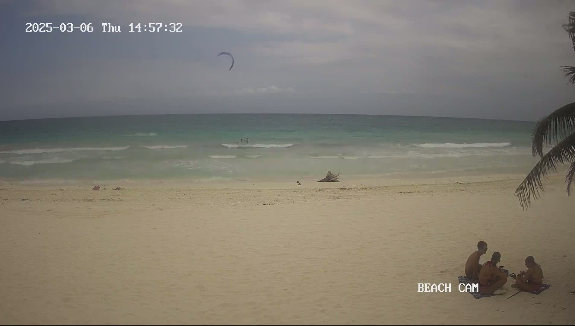 A sandy beach with a calm, light-green ocean under an overcast sky features a kite surfer in the distance and a small group of people sitting on the sand.