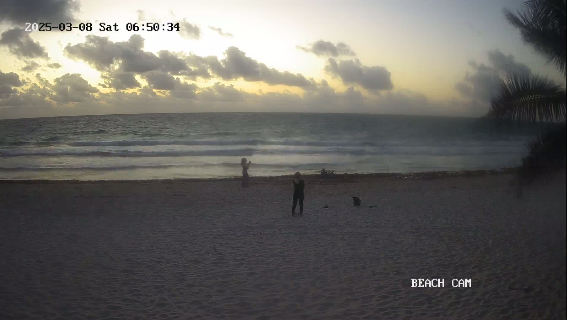 Several people stand on a sandy beach at sunrise under a partly cloudy sky, with gentle waves lapping the shore.