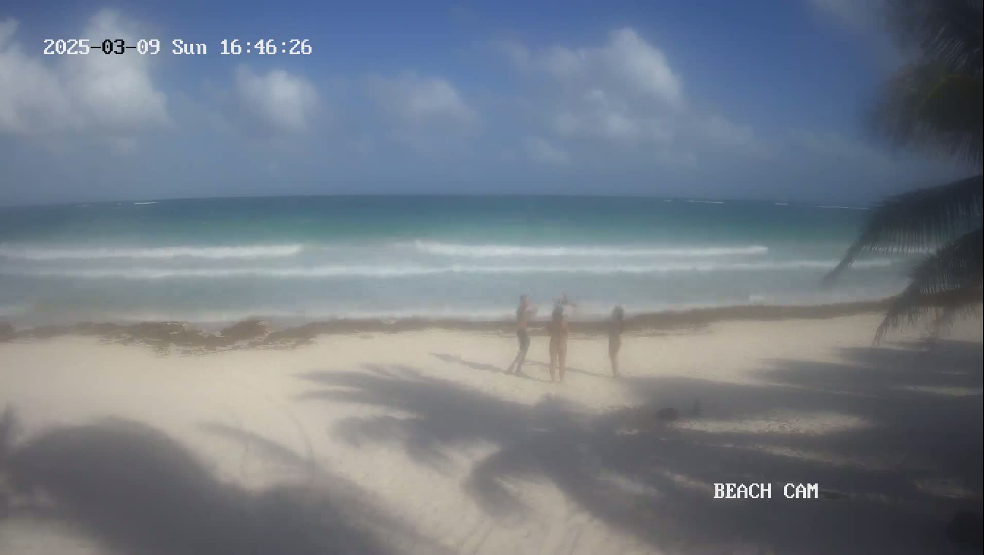 Three people stand on a white sand beach near a turquoise ocean with small waves under a partly cloudy sky.