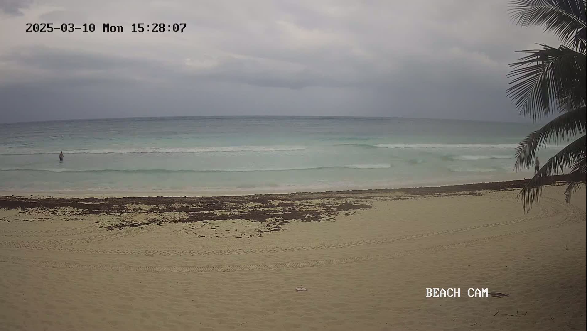 A person wades in the pale-turquoise ocean on a sandy beach under an overcast sky.