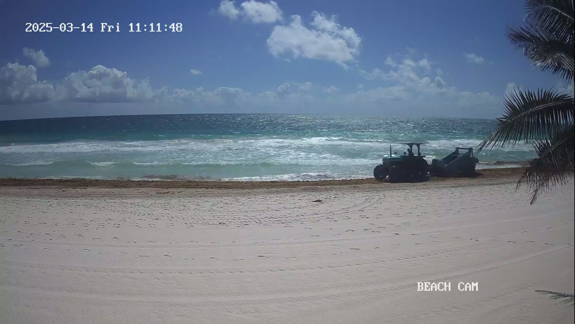 A tractor and attached beach cleaning equipment are on a white sand beach next to a teal ocean under a partly cloudy sky.
