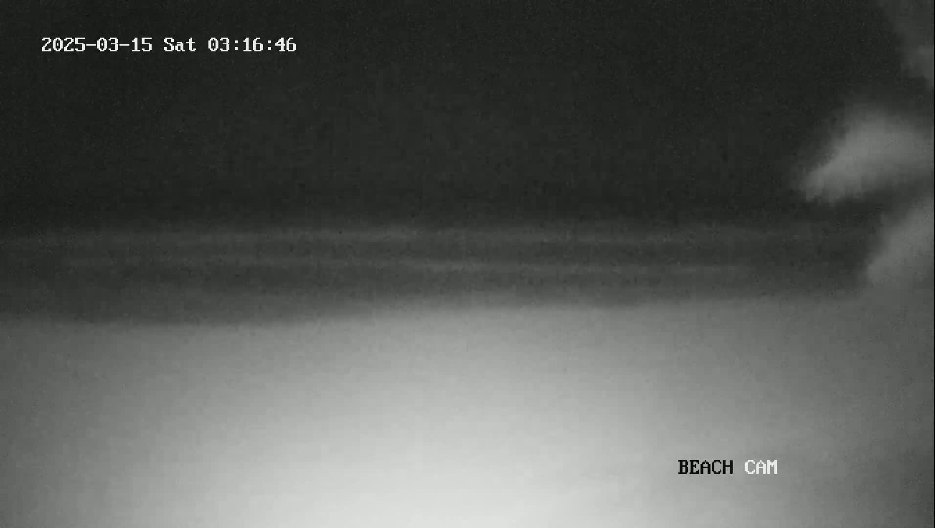 A low-light, nighttime beach scene shows dark clouds overhead, a lighter horizon line, and a mostly clear, dark sky.