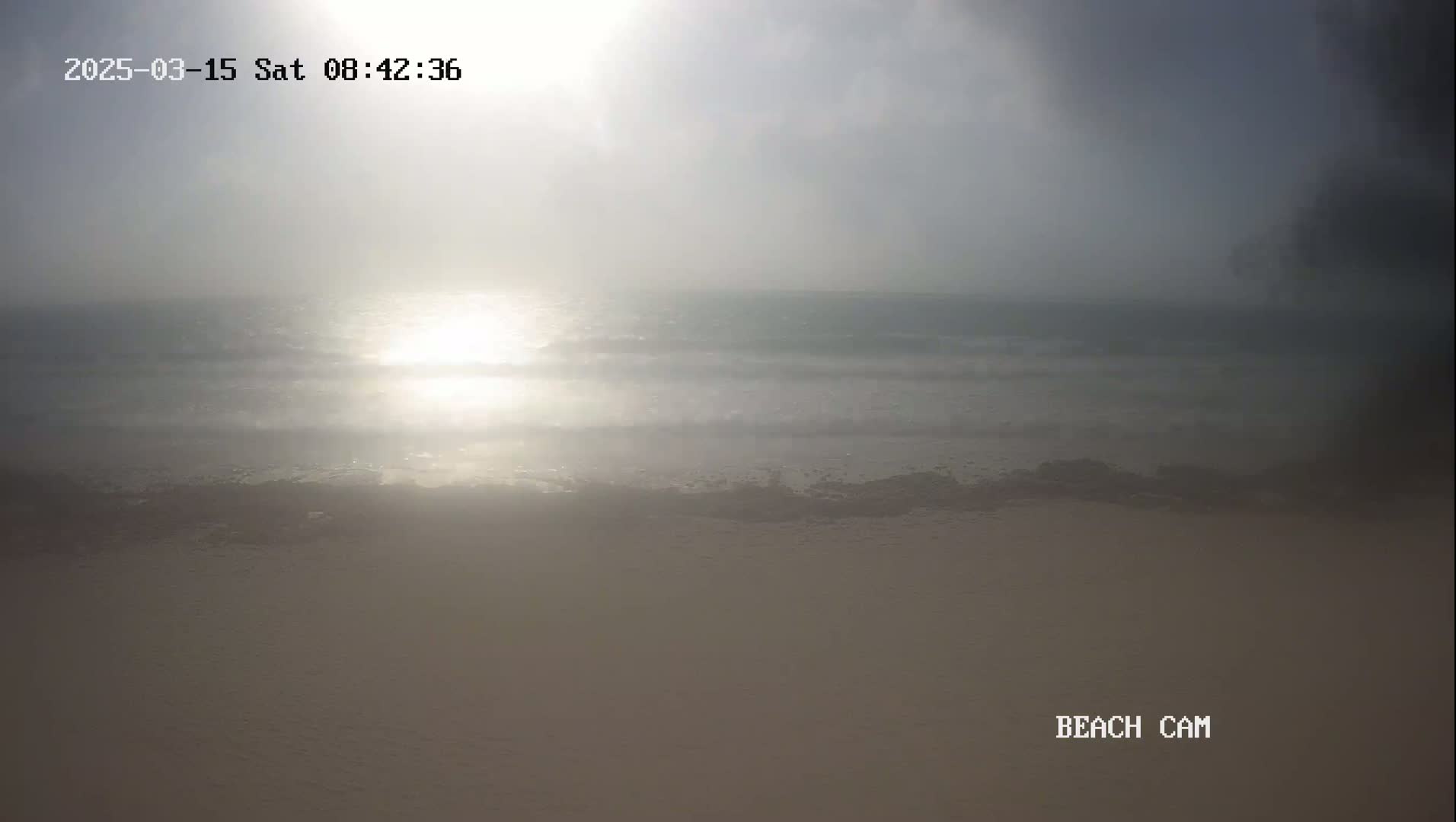 A hazy sunrise illuminates a calm ocean meeting a sandy beach.