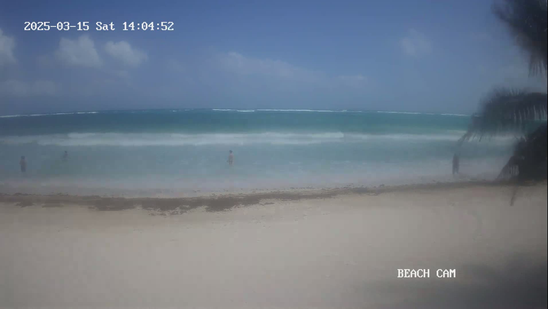 A mostly clear sky overlooks a beach with light waves and a few people wading in the shallow water.