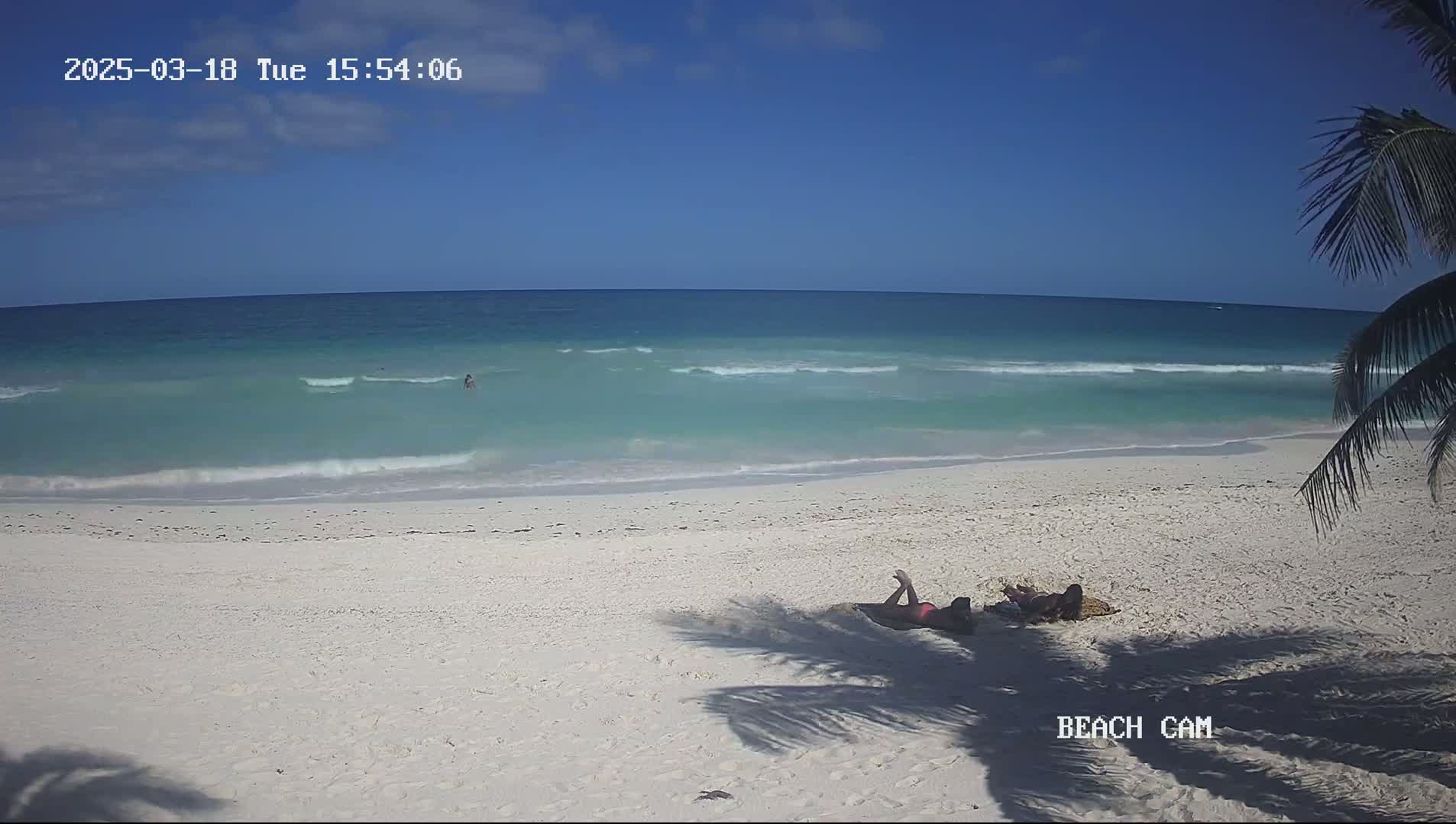 Two people lie on a white sandy beach next to a calm, light teal ocean under a clear blue sky.