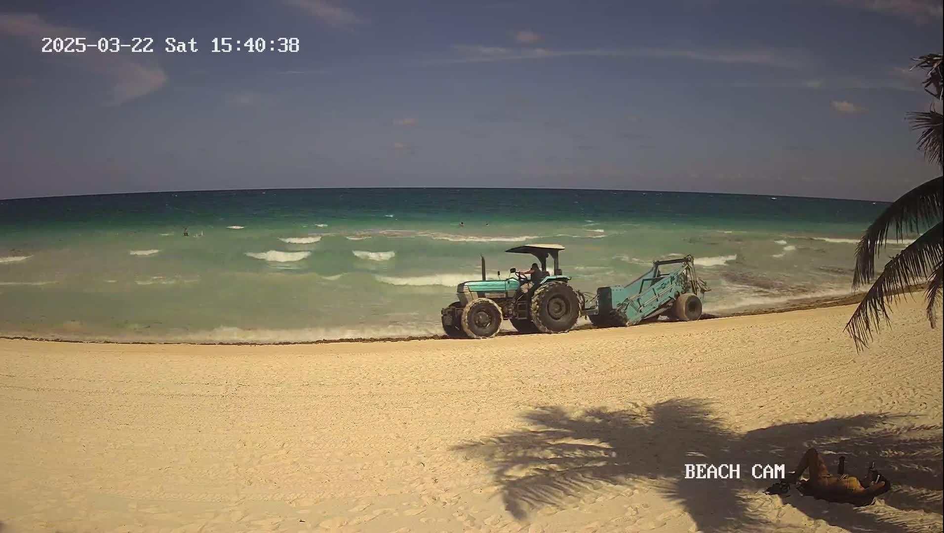 A teal tractor with an attached cleaning implement is on a sandy beach next to a turquoise ocean with small waves under a mostly clear sky.