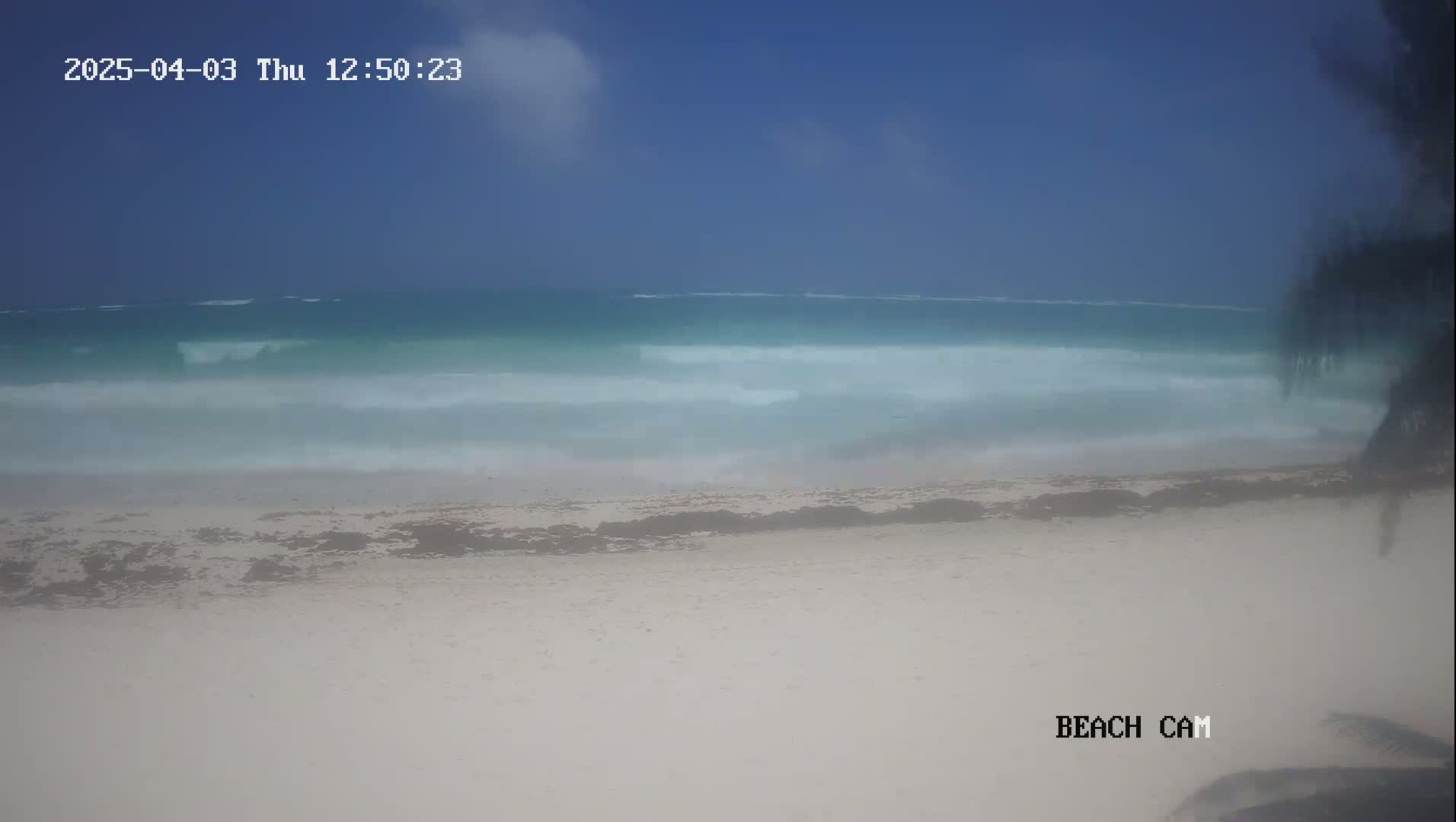 A mostly clear, hazy day shows a light-blue ocean with small waves gently lapping a white sandy beach.