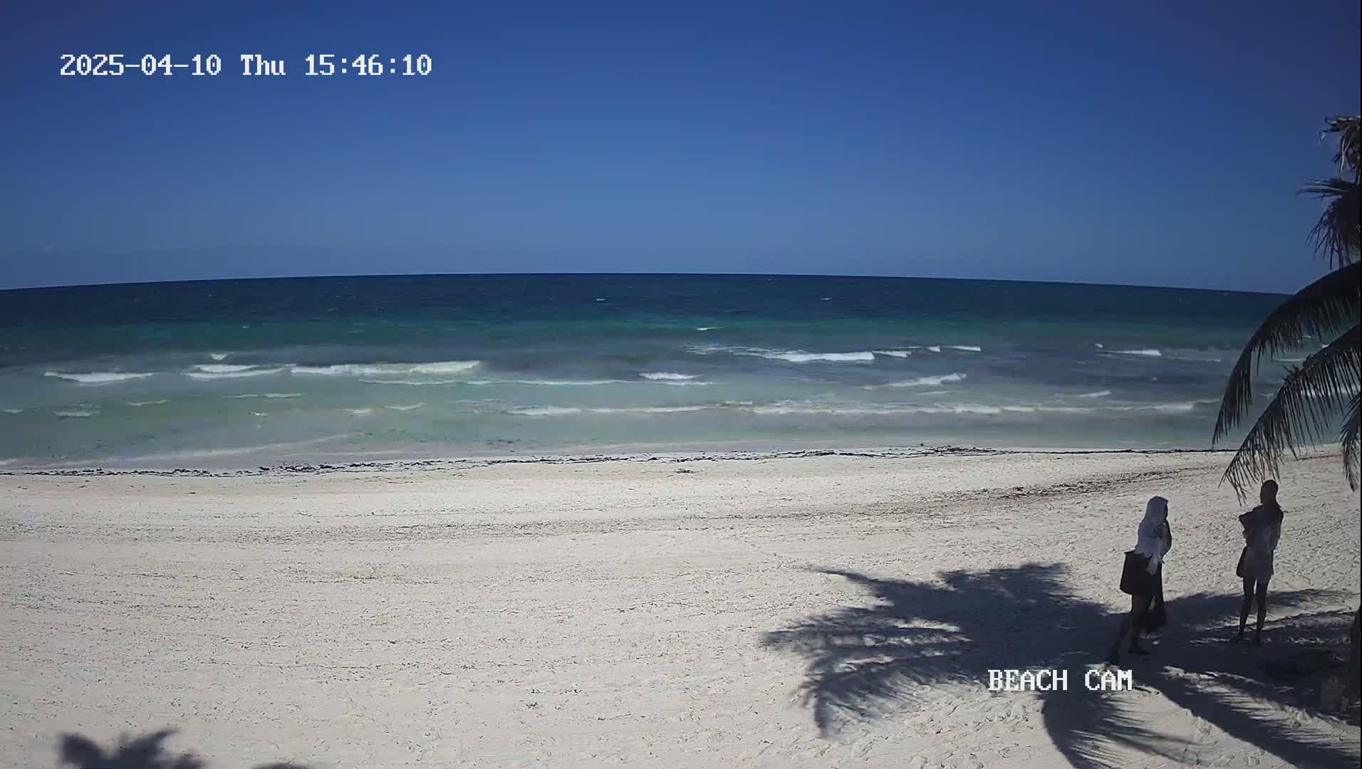 Two people stand on a white sand beach near turquoise water with small waves under a clear blue sky.