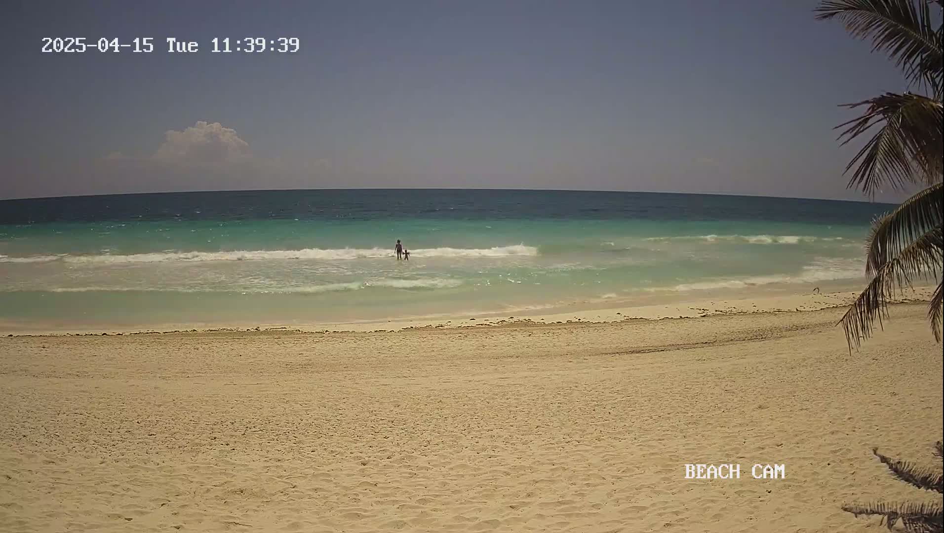 A sandy beach with calm, light turquoise waves and two people wading in the shallows under a mostly clear sky.
