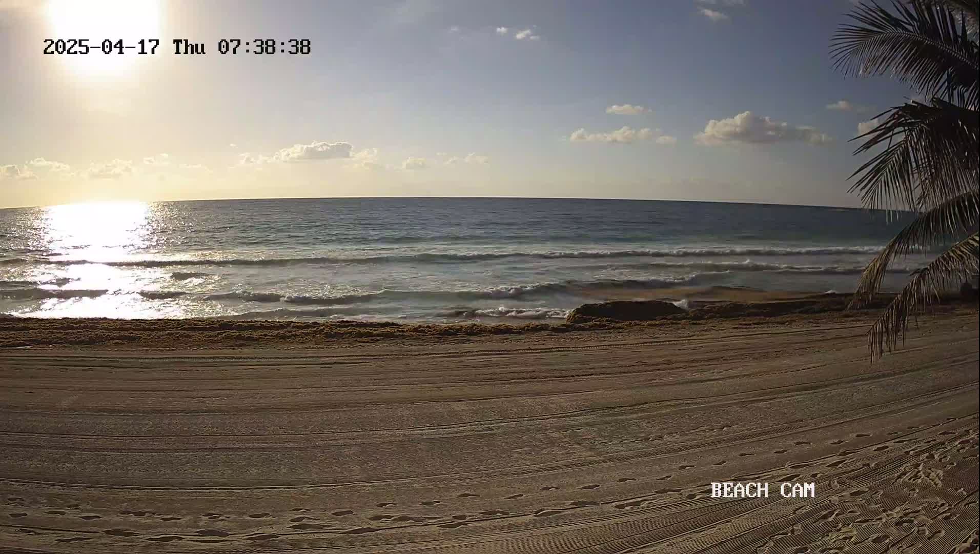 A sandy beach with footprints shows a calm ocean under a partly cloudy sunrise.