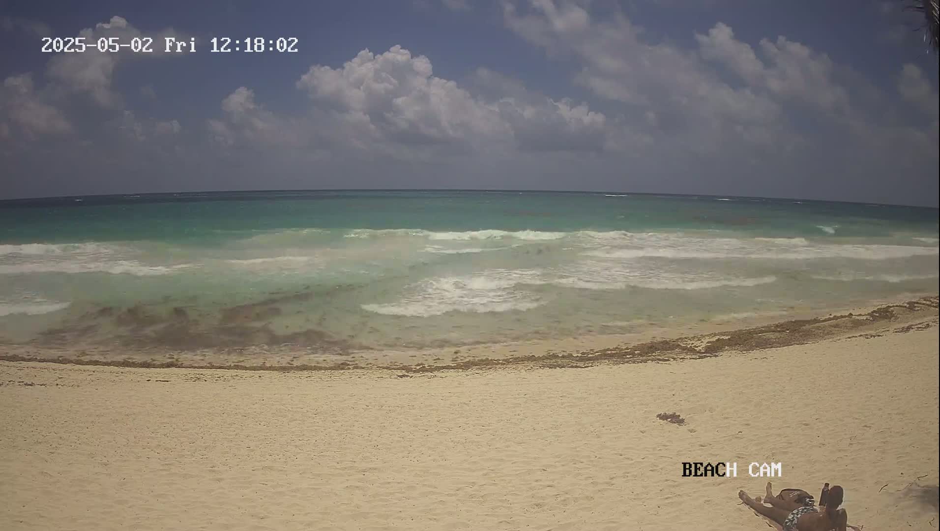 A partly cloudy day shows a sandy beach with light waves lapping the shore, and two people lying on the sand.