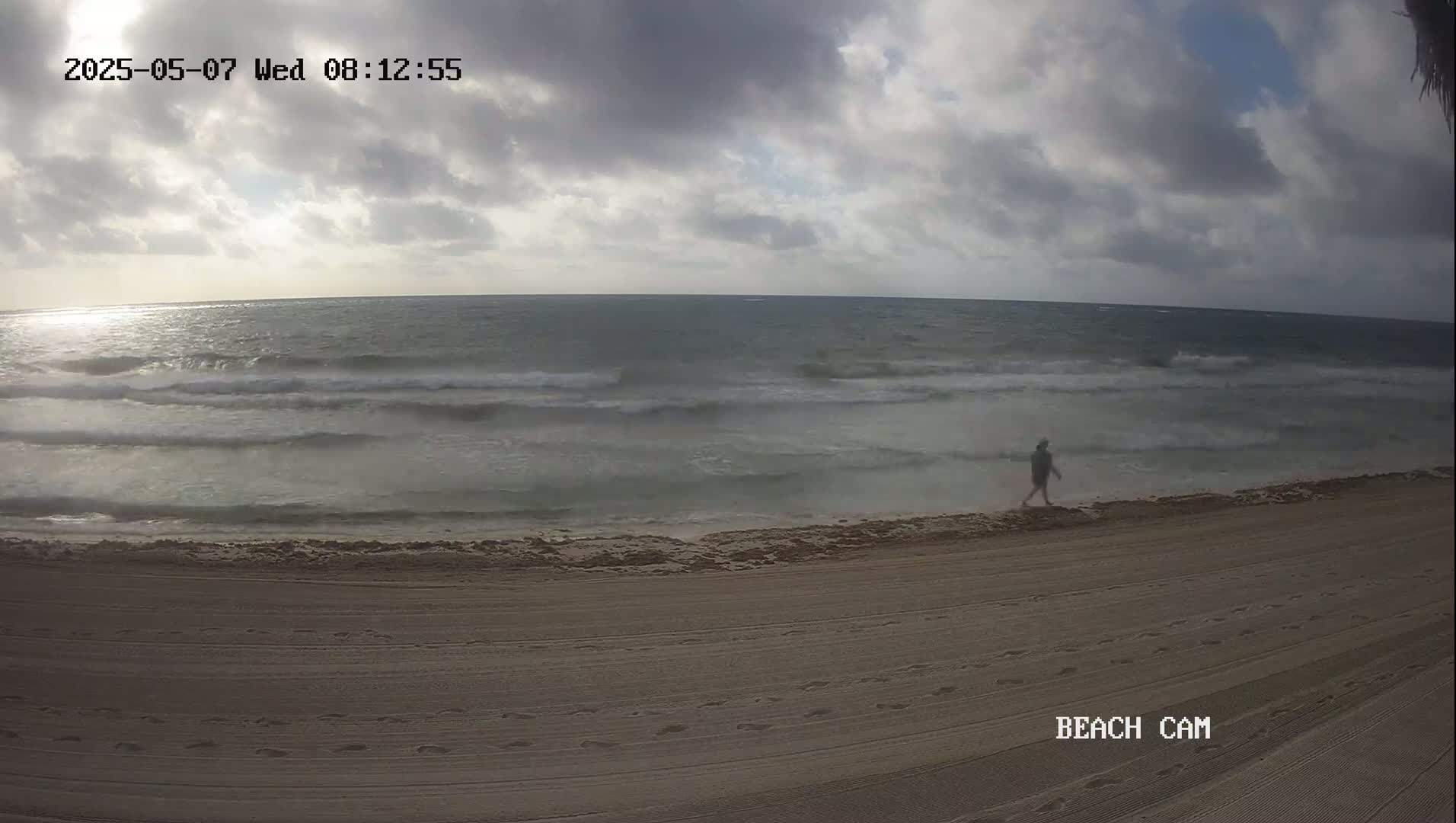 A person walks along a sandy beach under a cloudy sky with somewhat choppy waves.