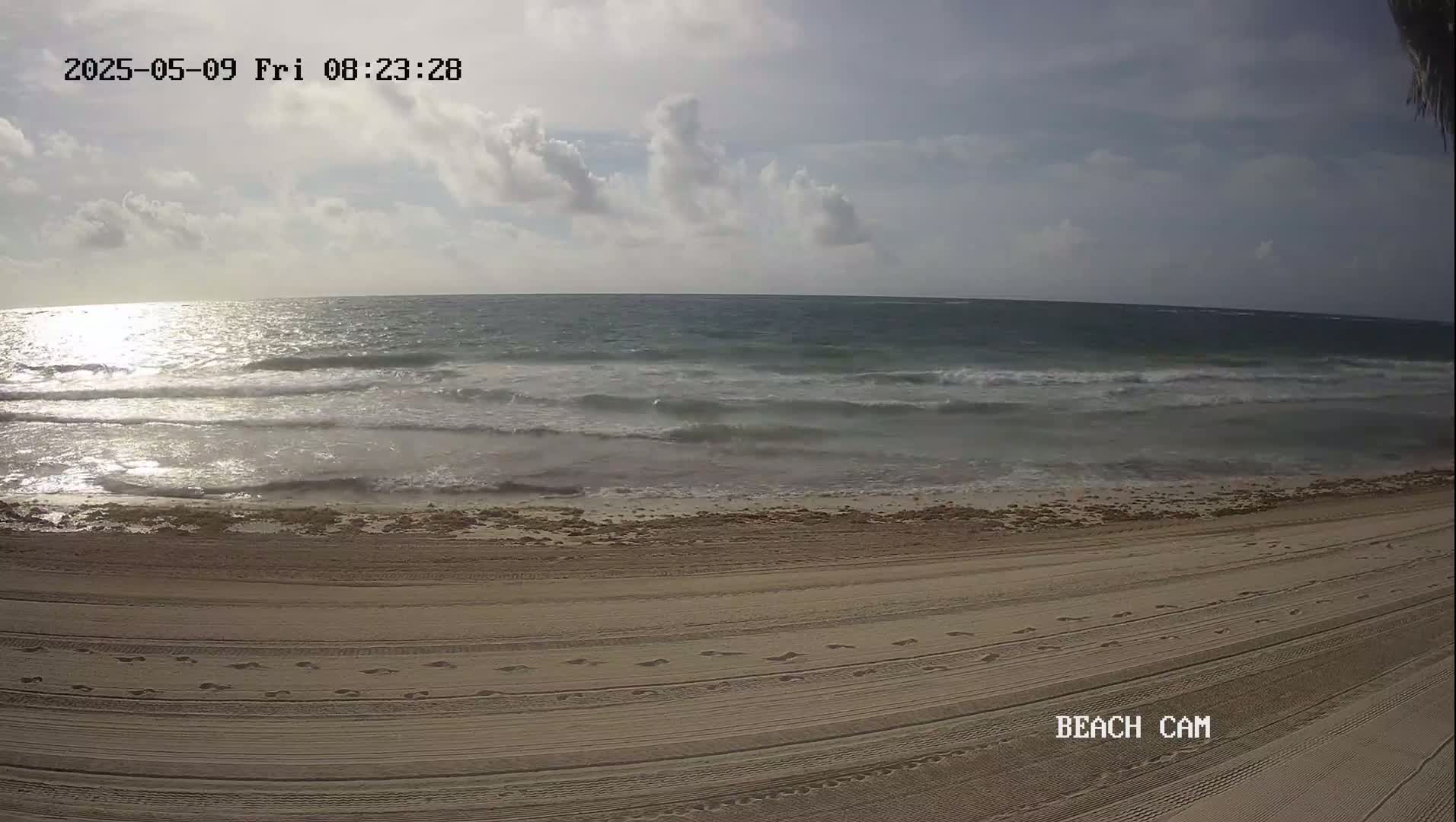 A sandy beach with footprints shows moderate waves lapping the shore under a partly cloudy sky.
