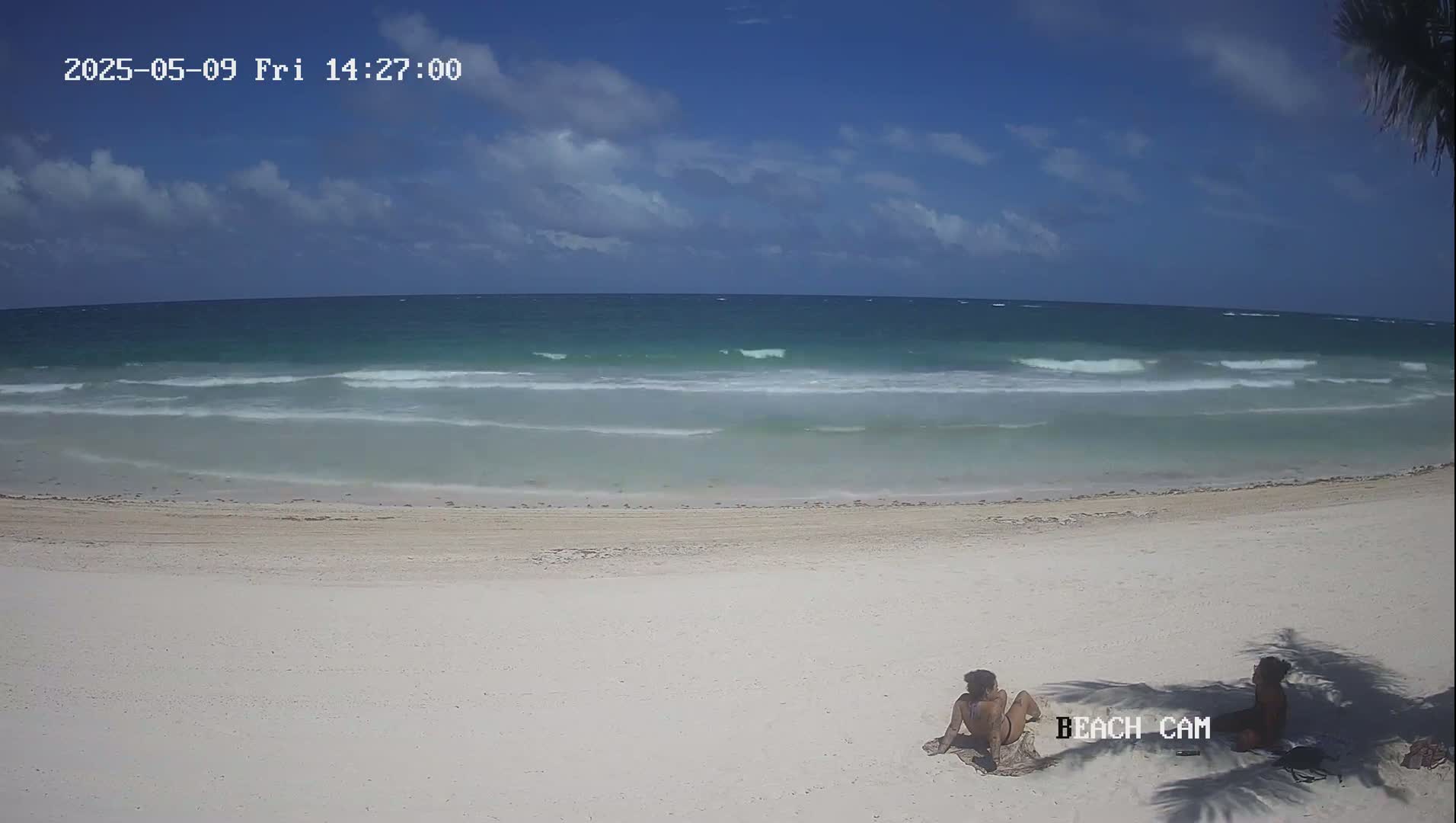 Two people sit on a white sand beach overlooking a teal ocean with small waves under a partly cloudy sky.