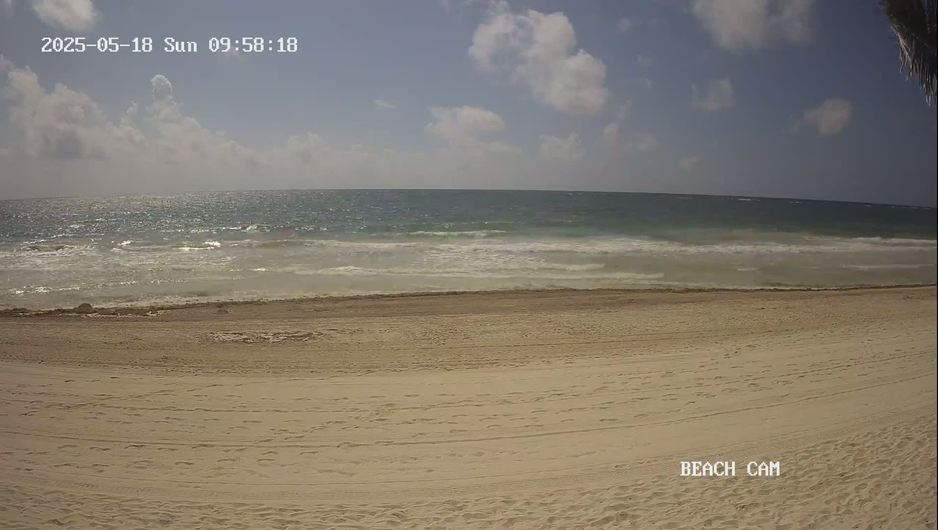 A sandy beach with footprints stretches towards a moderately wavy ocean under a partly cloudy sky.